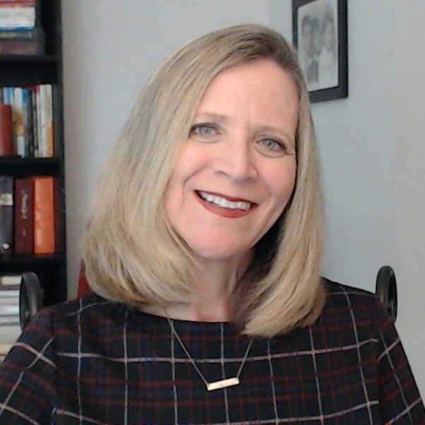 A smiling woman with shoulder-length blonde hair sitting in a room with bookshelves and framed pictures.