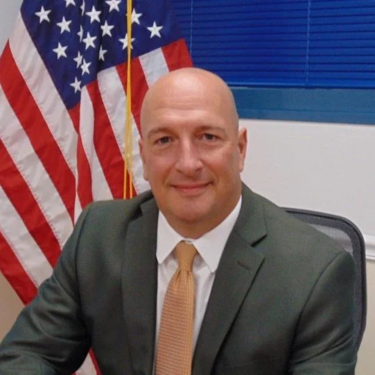 A man in a suit sitting at a desk with an American flag in the background.