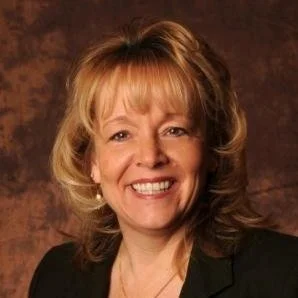 Smiling woman with blonde, curly hair and a dark blazer against a brown backdrop.