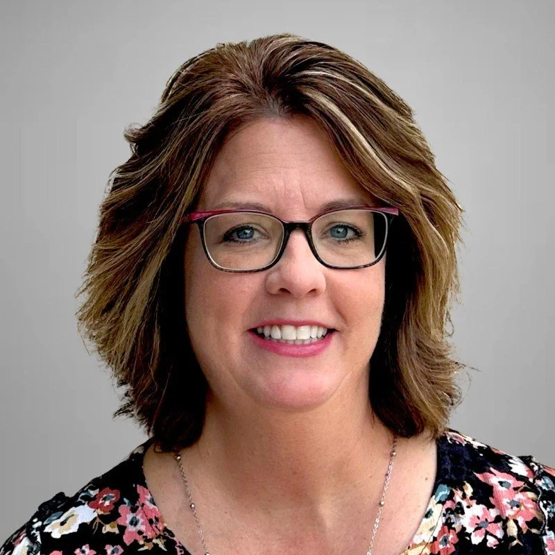 A woman with shoulder-length brown hair wearing glasses, smiling, dressed in a floral top, against a plain gray background.