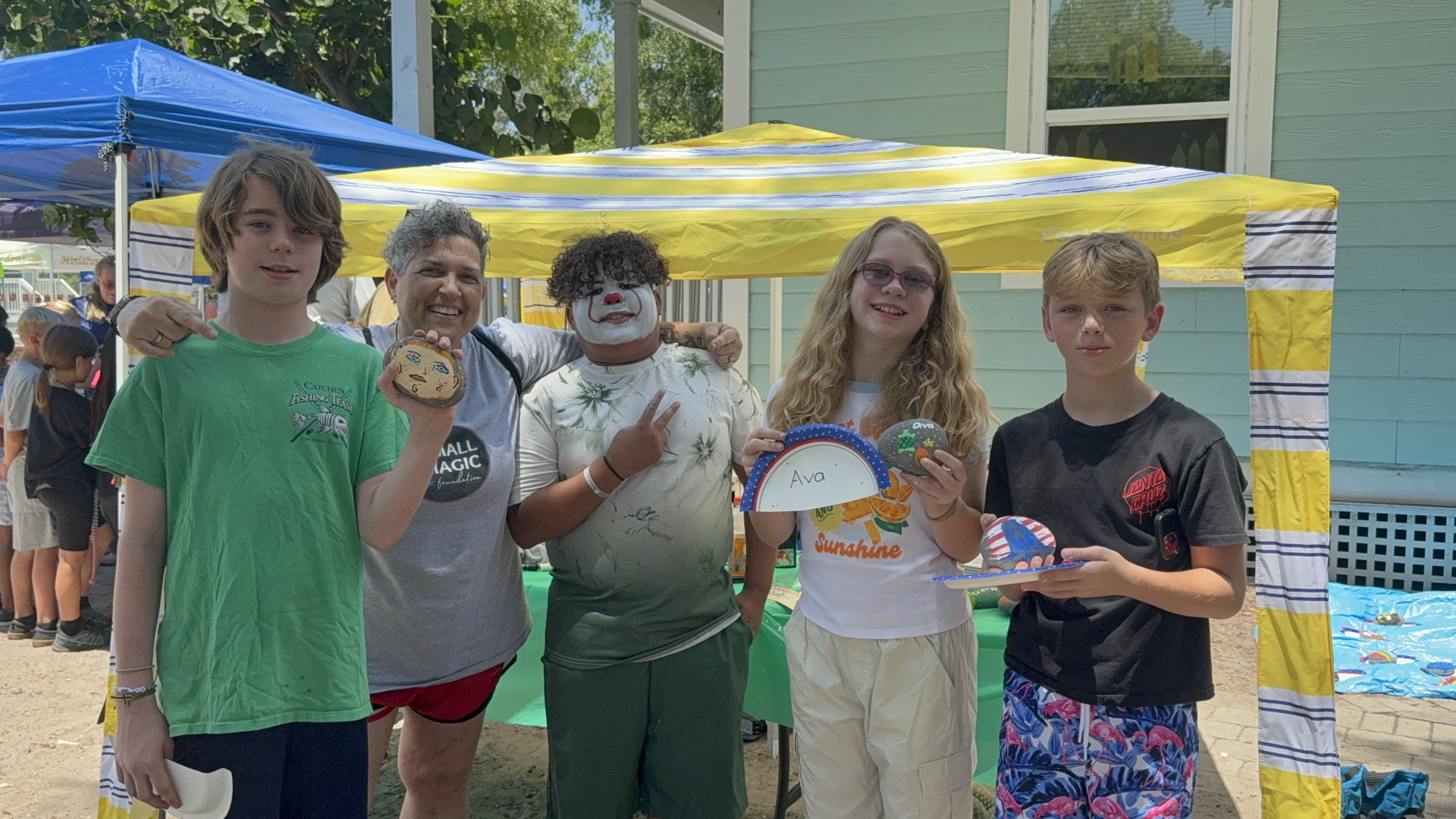Group of five kids and an adult with clown makeup standing under a yellow and blue tent at an outdoor event, holding painted rocks and a paper plate with the name "Ava" written on it