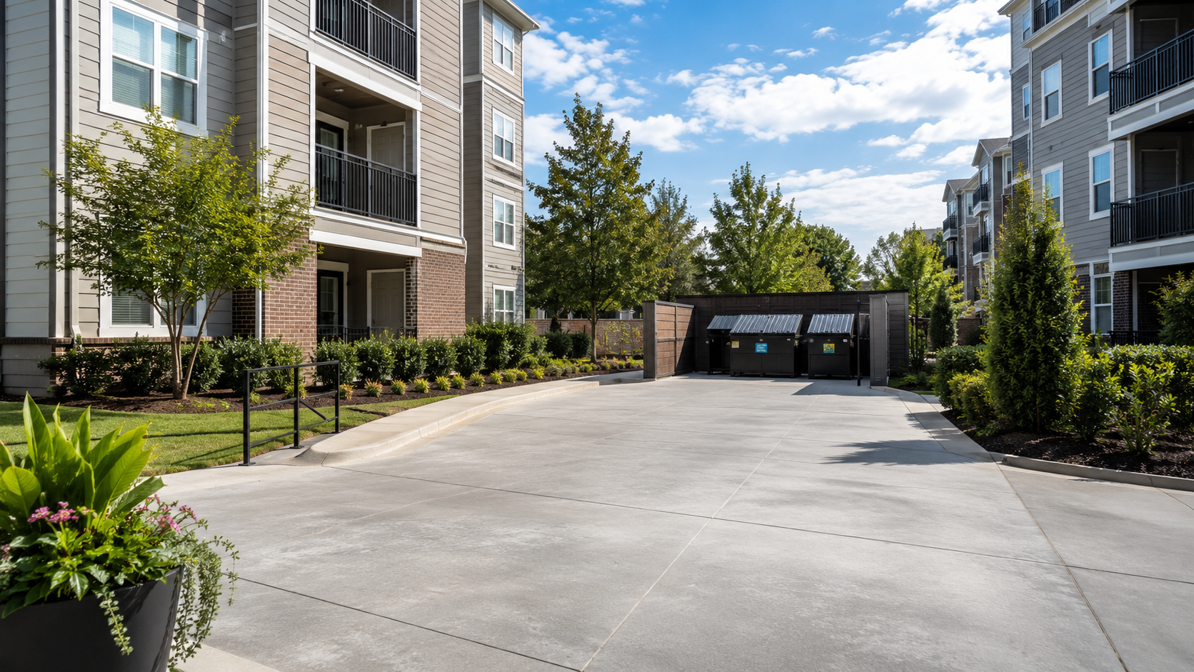 A clean courtyard area in an apartment complex with landscaped bushes and trees, surrounded by multi-story buildings, and a trash enclosure with dumpsters against a bright blue sky with scattered clouds.