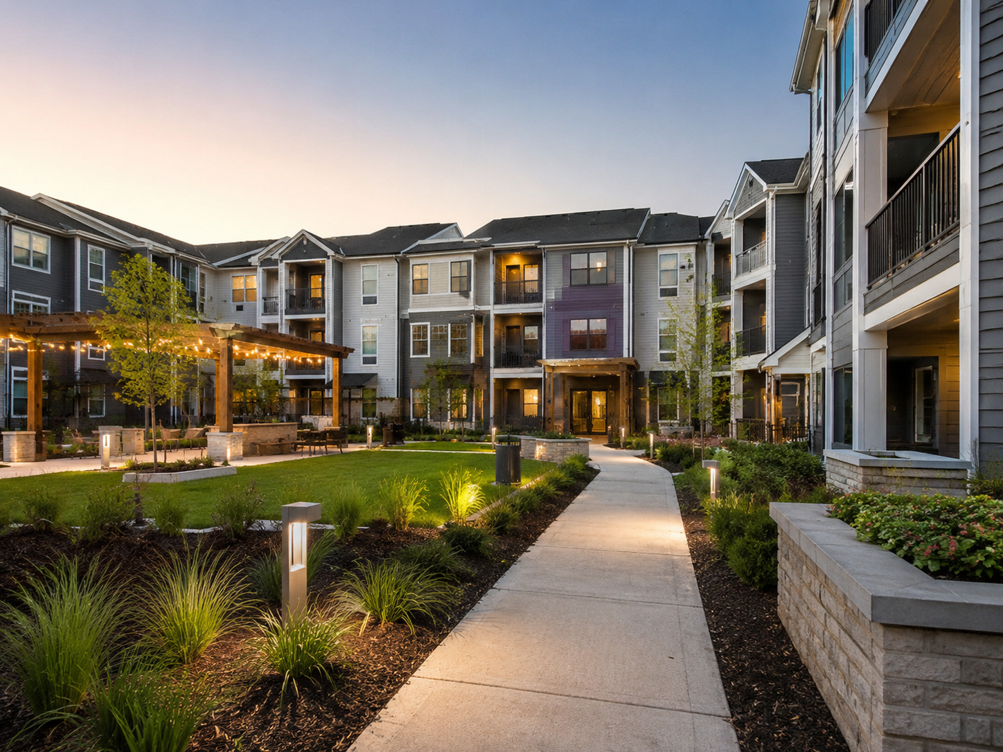 View of an apartment complex courtyard at dusk with a paved walkway, lush greenery, garden beds, and a shaded seating area with string lights.