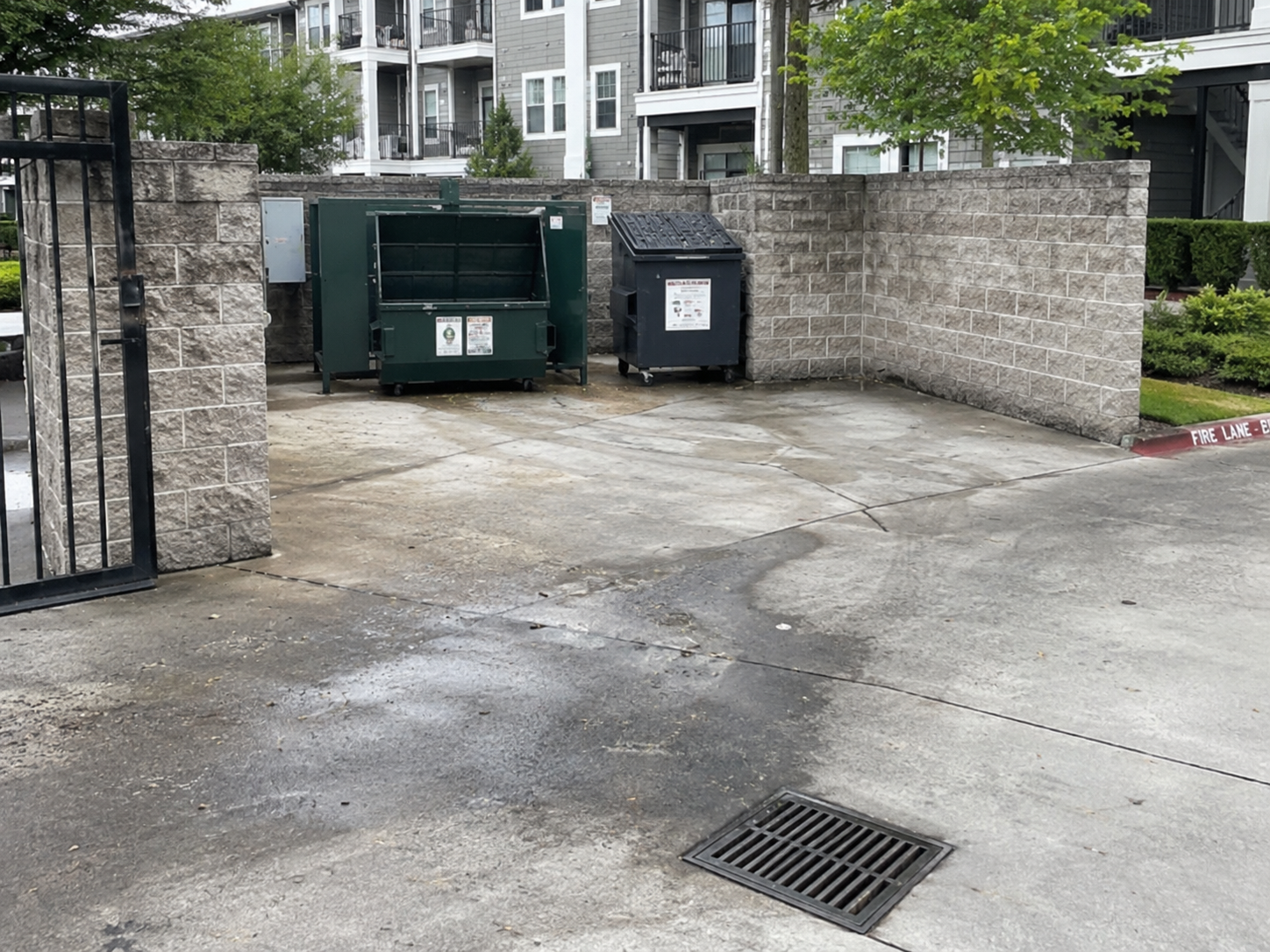 Apartment complex courtyard with waste disposal bins and a brick wall, wet concrete ground, and greenery in the background.
