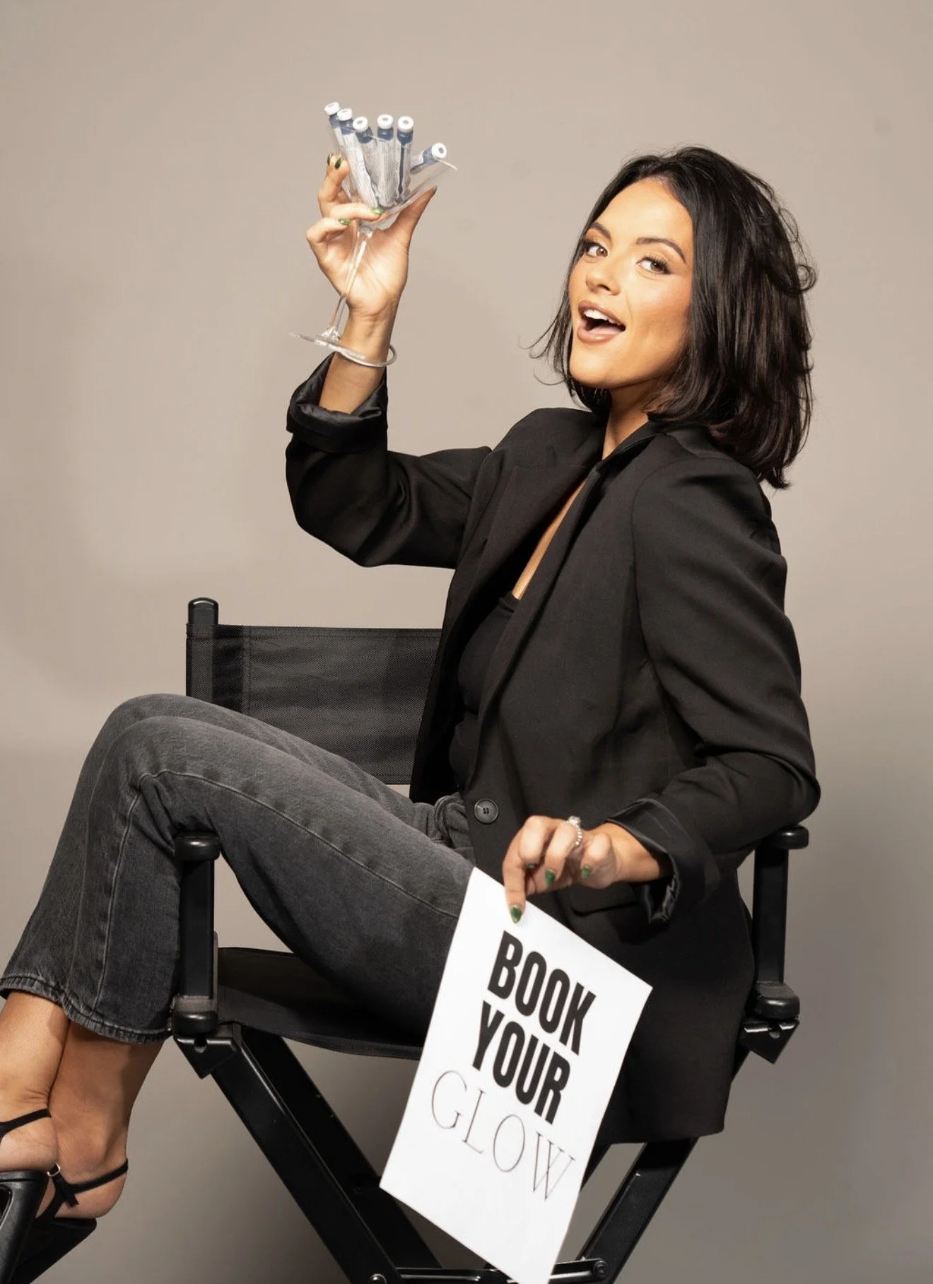 A woman with dark, wavy hair and makeup, wearing a black blazer and grey jeans, sitting on a director's chair, holding a small mirror or compact, with a sign that says 'BOOK YOUR GLOW' hanging from her lap.