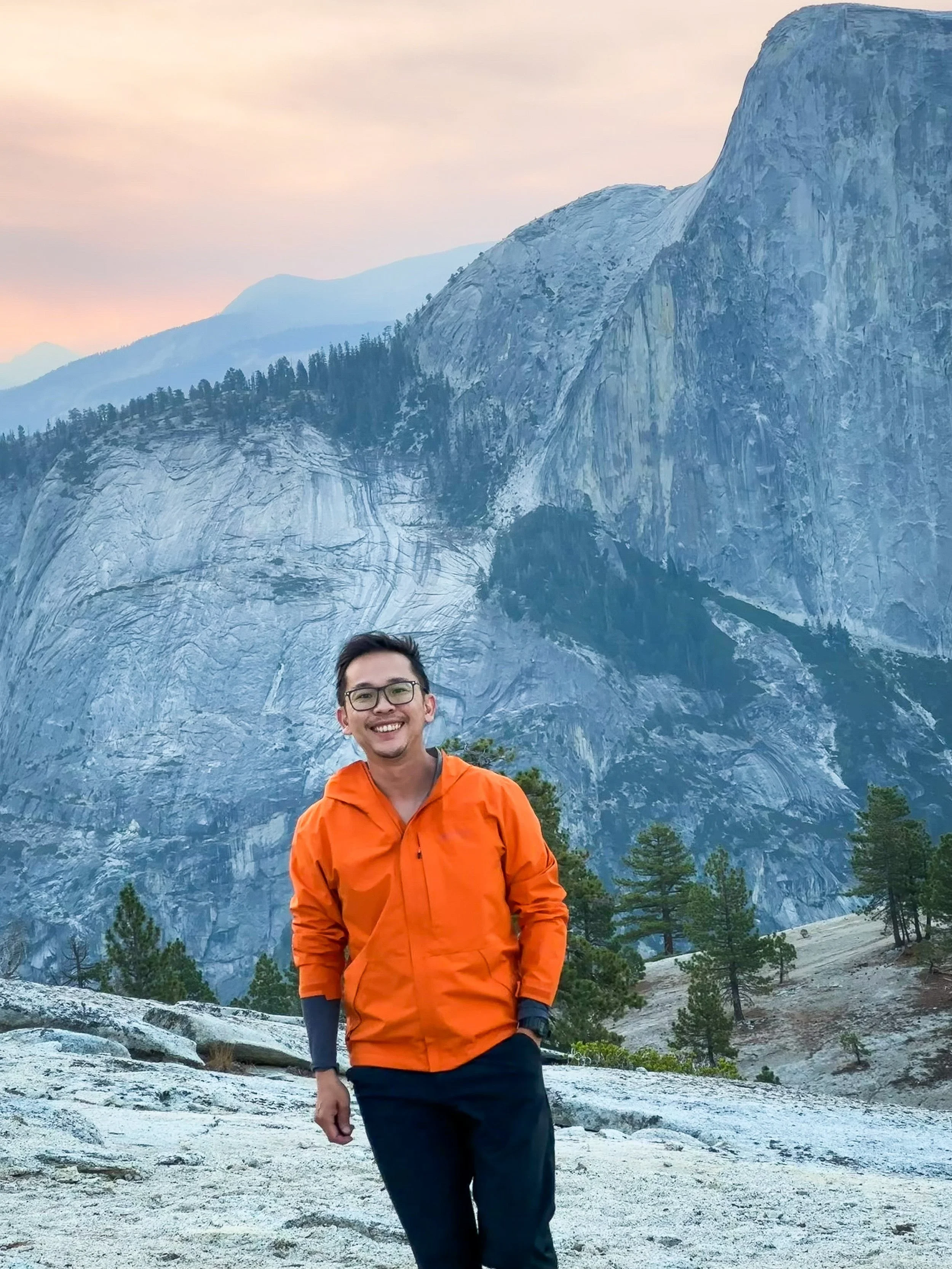 A smiling man wearing glasses, an orange jacket, and dark pants standing on rocky terrain in front of a mountain landscape with tall granite cliffs and pine trees, under a pink and blue sky.