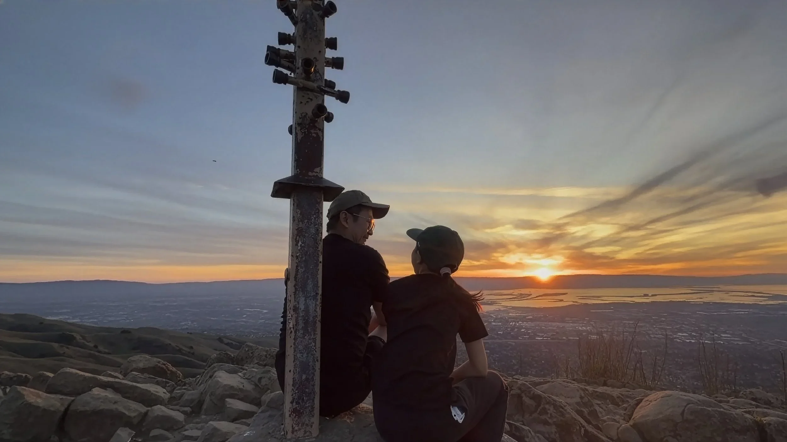 Two people sitting on rocks near a weathered metal pole with multiple cameras, watching a sunset over a body of water. One person is an older man wearing a cap, and the other is a younger person with a ponytail and a cap, both facing the sunset.