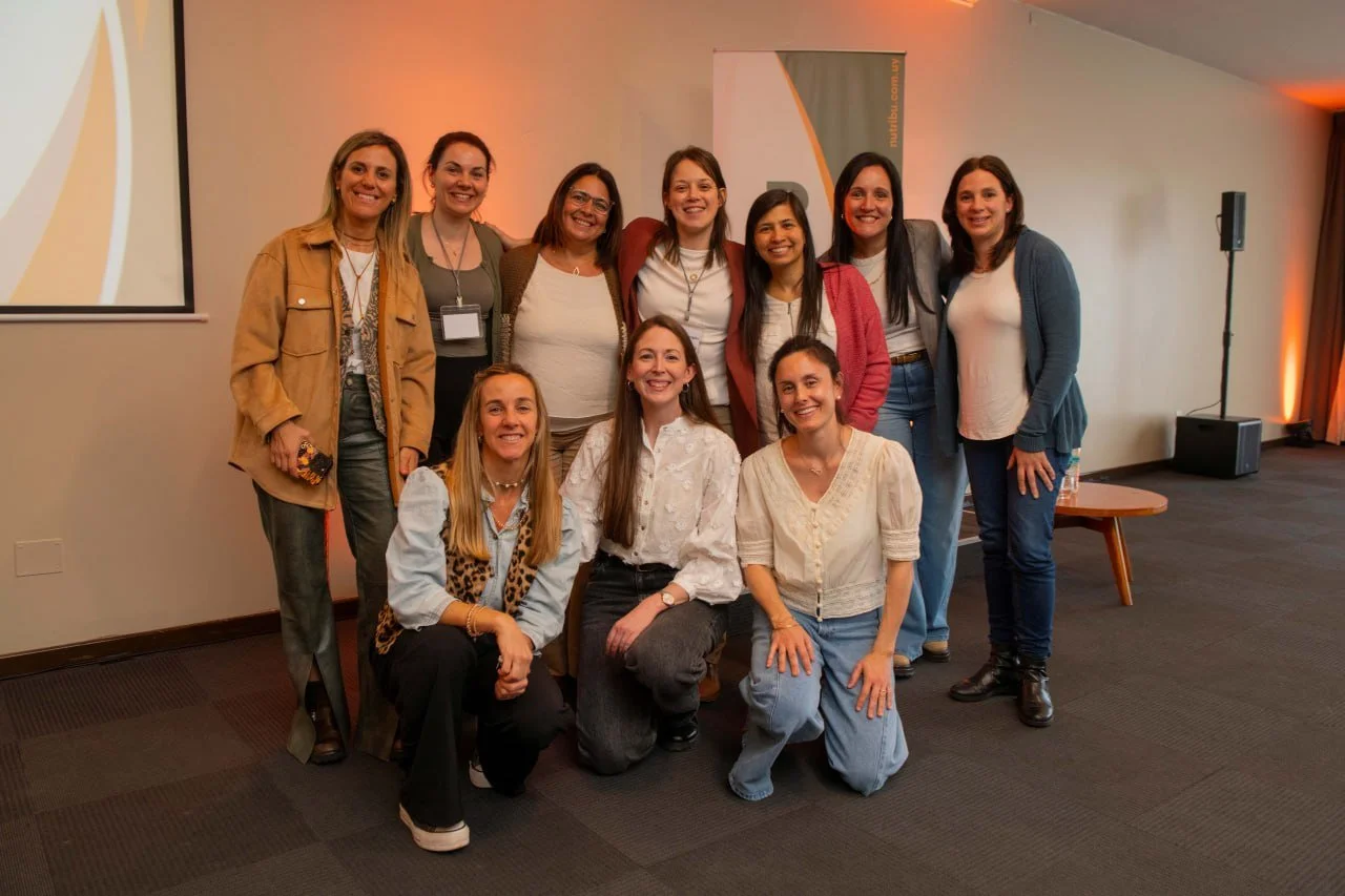 Grupo de diez mujeres sonriendo en una conferencia o evento, posando para la foto en un espacio interior con equipo de sonido y pantalla de proyección.