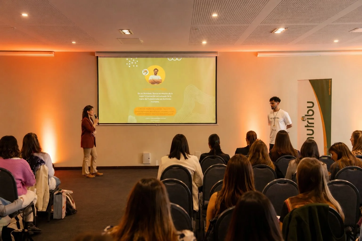Una presentación en un salón con varias personas sentadas, dos oradores en el frente, uno con micrófono, y una pantalla con una diapositiva. Hay una pancarta a la derecha con el logo y el nombre de la organización.