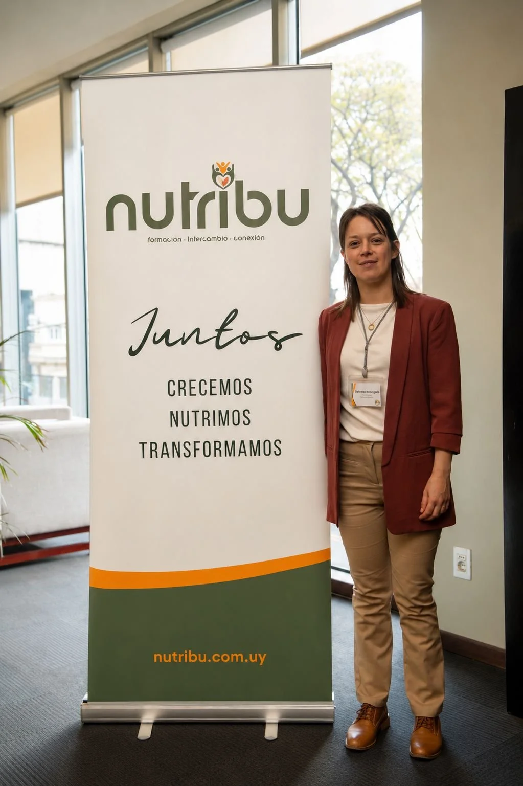 Mujer de pie junto a una pancarta que dice 'Juntos' y tiene los siguientes textos: 'Creamos, Nutrimos, Transformamos' y la página web 'ntribu.com.uy'.