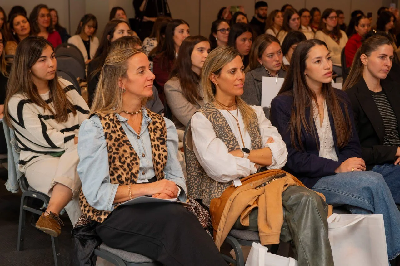 Grupo de mujeres jóvenes y adultas asistiendo a una conferencia, sentadas en silencio y prestando atención.