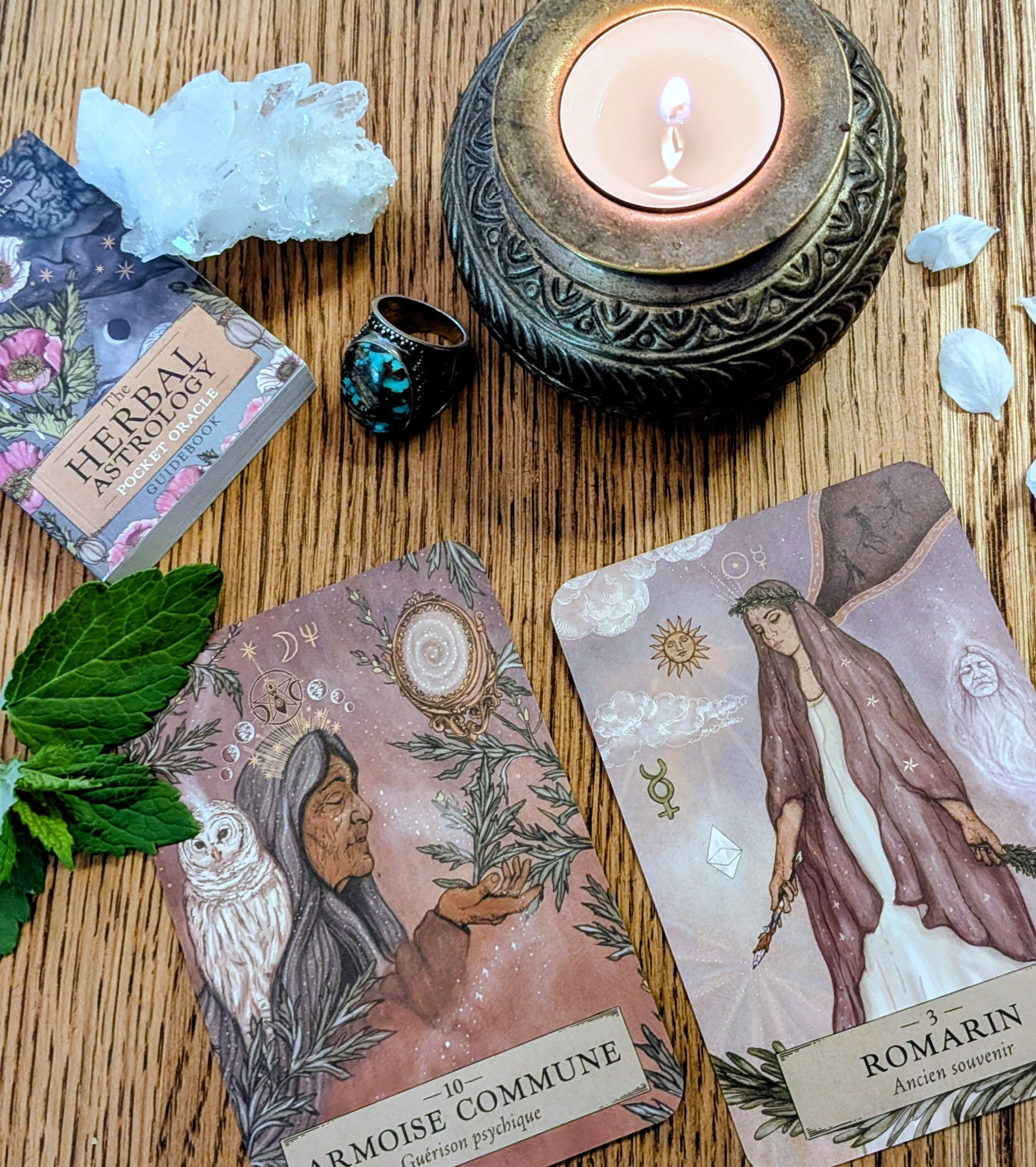 A lit candle in a decorative holder, a turquoise ring, a bundle of white crystals, a tarot card, a stack of tarot cards, and a sprig of fresh mint leaves arranged on a wooden surface.