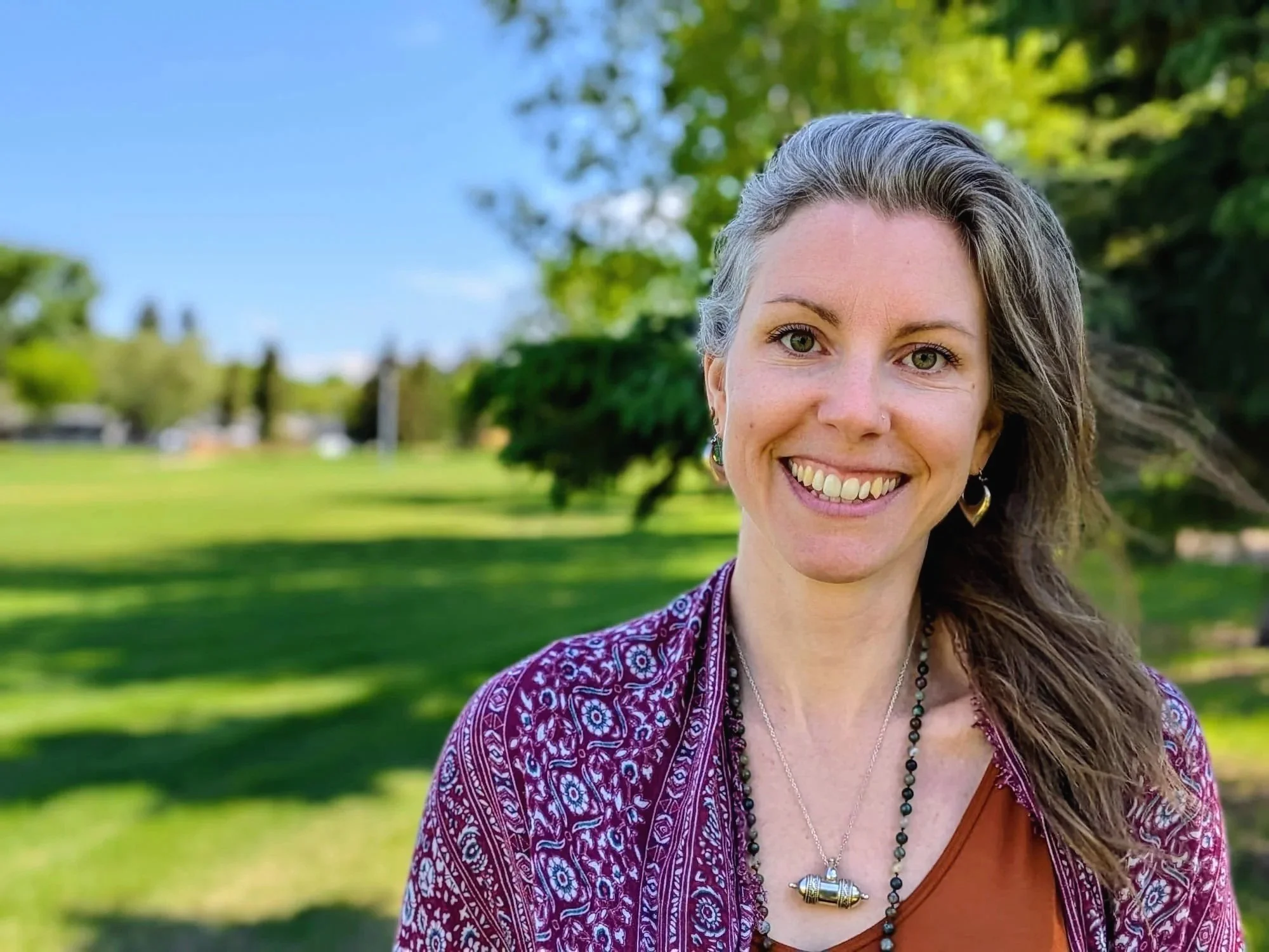 Close-up of a smiling woman with long, gray and brown hair, wearing earrings and layered necklaces, outdoors with green trees and a blue sky in the background.