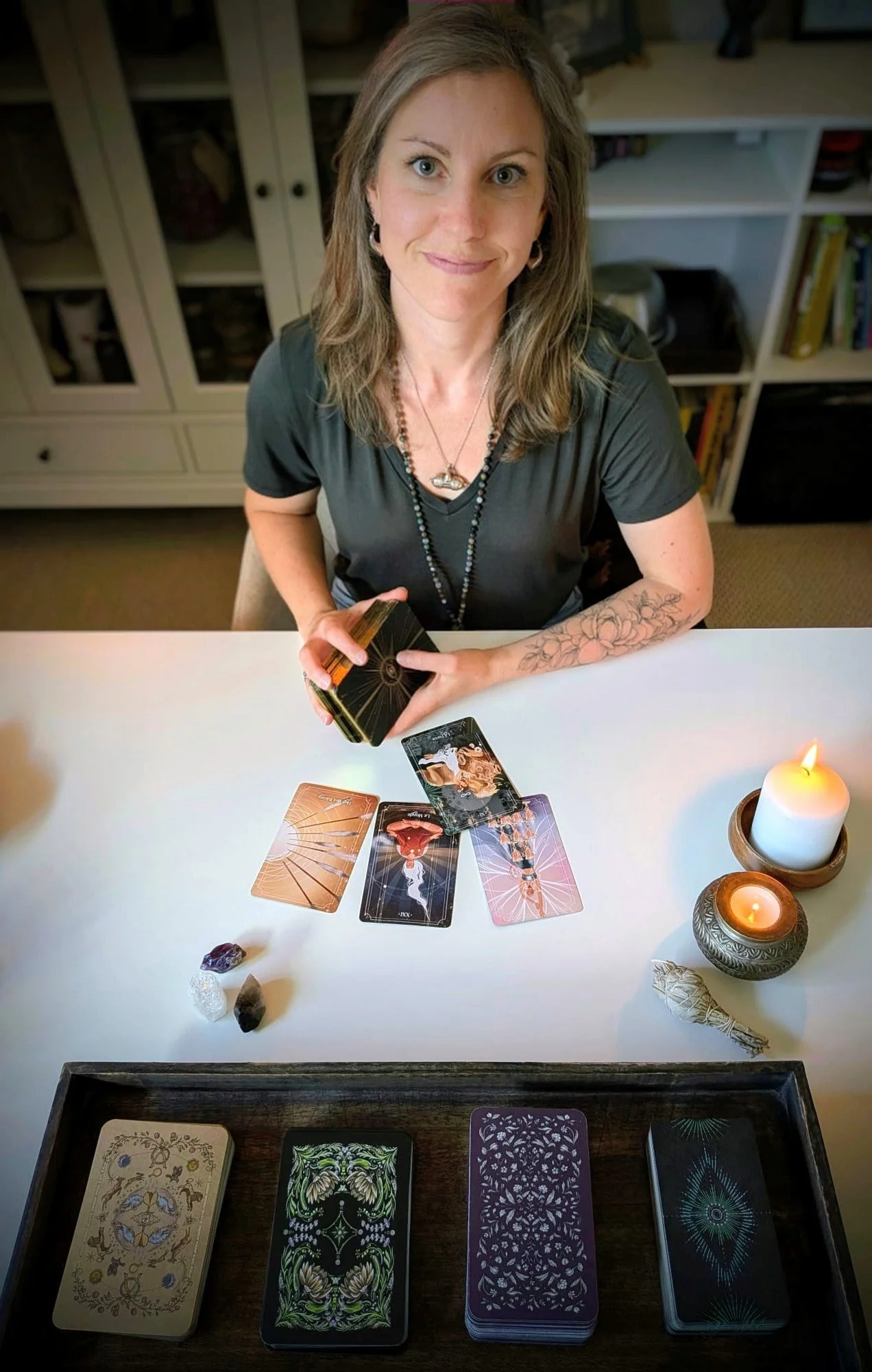 A woman with long hair, wearing a black shirt and jewelry, is sitting at a white table with tarot cards, crystals, candles, and a tray of additional cards for a tarot reading.