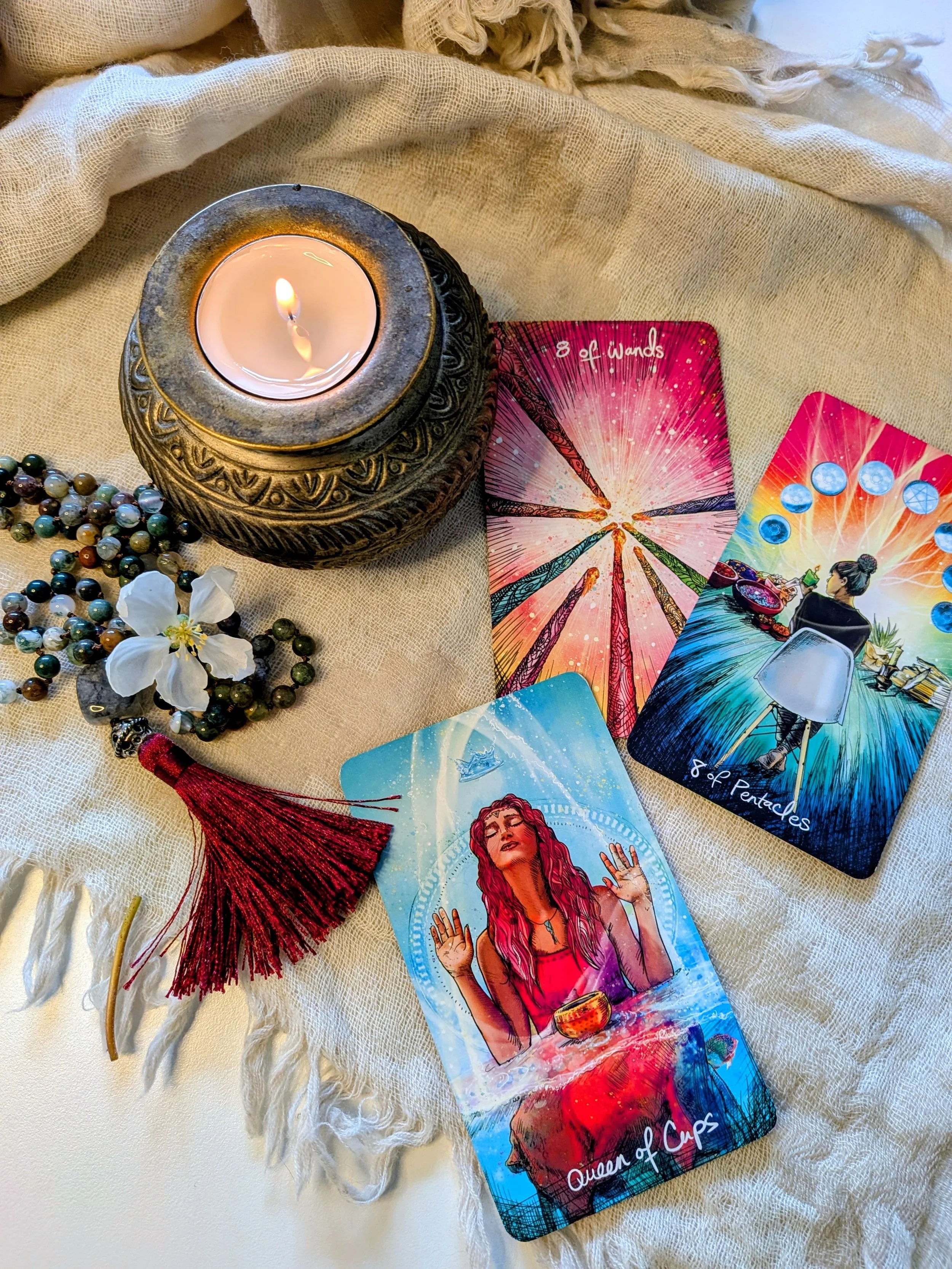 A lit candle in an ornate black holder, prayer beads, white flower, tarot cards, and a red tassel on a beige cloth surface.
