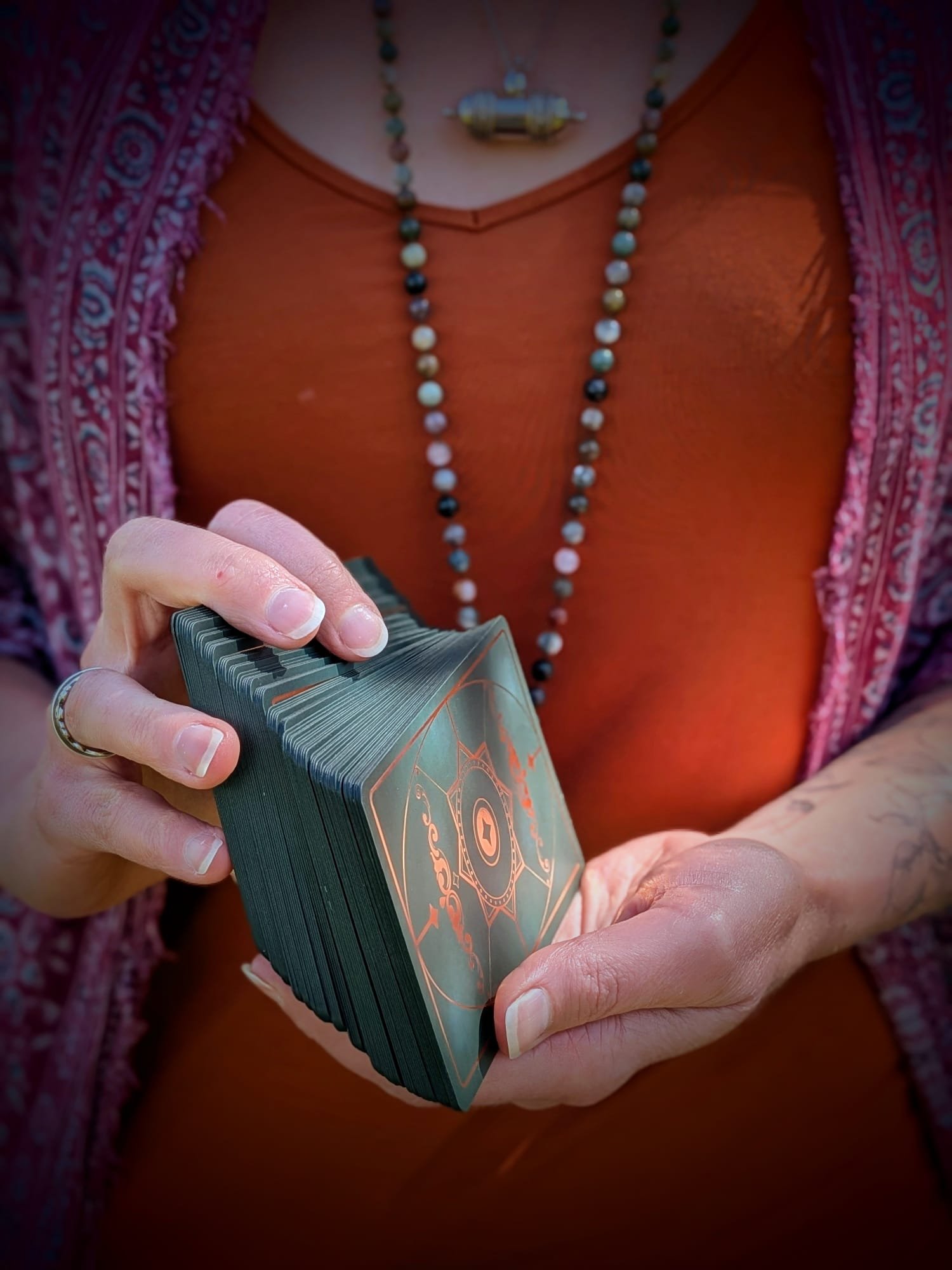 Close-up of a person holding a deck of tarot cards, with the focus on the cards and hands. The person is wearing an orange shirt, a pink patterned shawl, and a beaded necklace, with a metallic pendant.