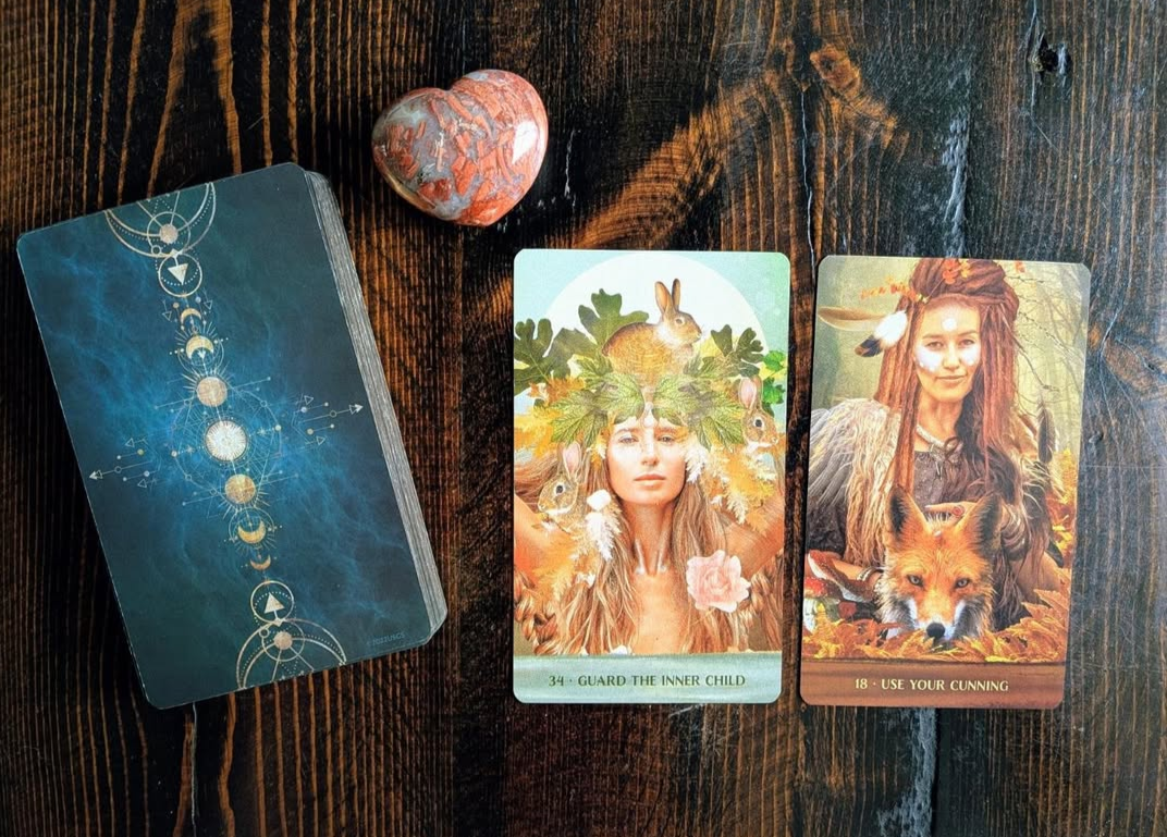 A set of tarot or oracle cards on a dark wooden surface, featuring two illustrated cards with women and animals, a small heart-shaped stone, and a card back with geometric symbols.