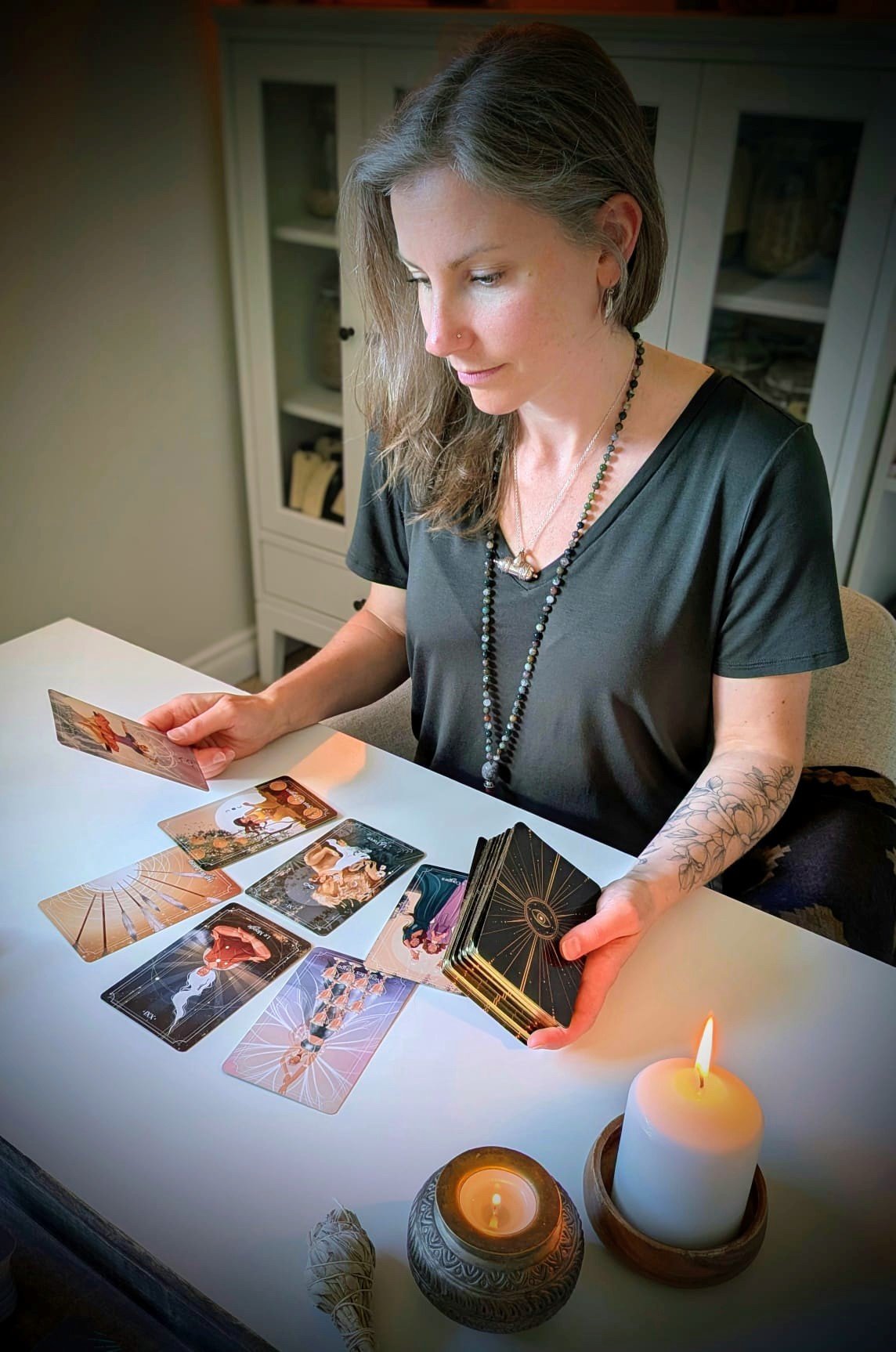 A woman with long, gray hair, wearing a black shirt and necklaces, sits at a white table with tarot cards spread out. She is holding some cards in one hand and a deck in the other. There are lit candles on the table and a cabinet with glass doors in the background.
