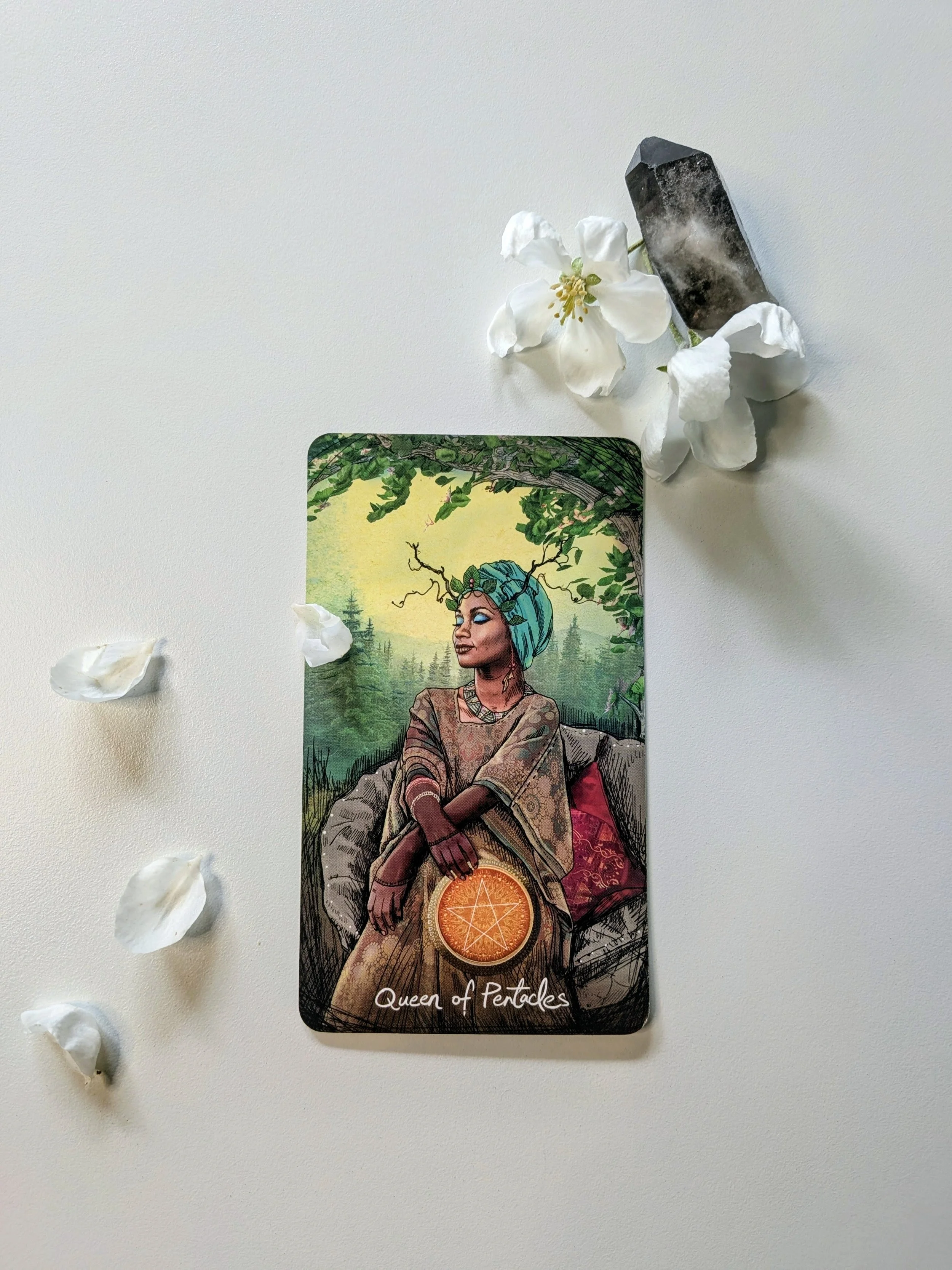 A tarot card titled Queen of Pentacles with an illustration of a woman sitting outdoors with greenery, a forest background, and a celestial sphere at her lap, surrounded by white flower petals and a black crystal on a white surface.