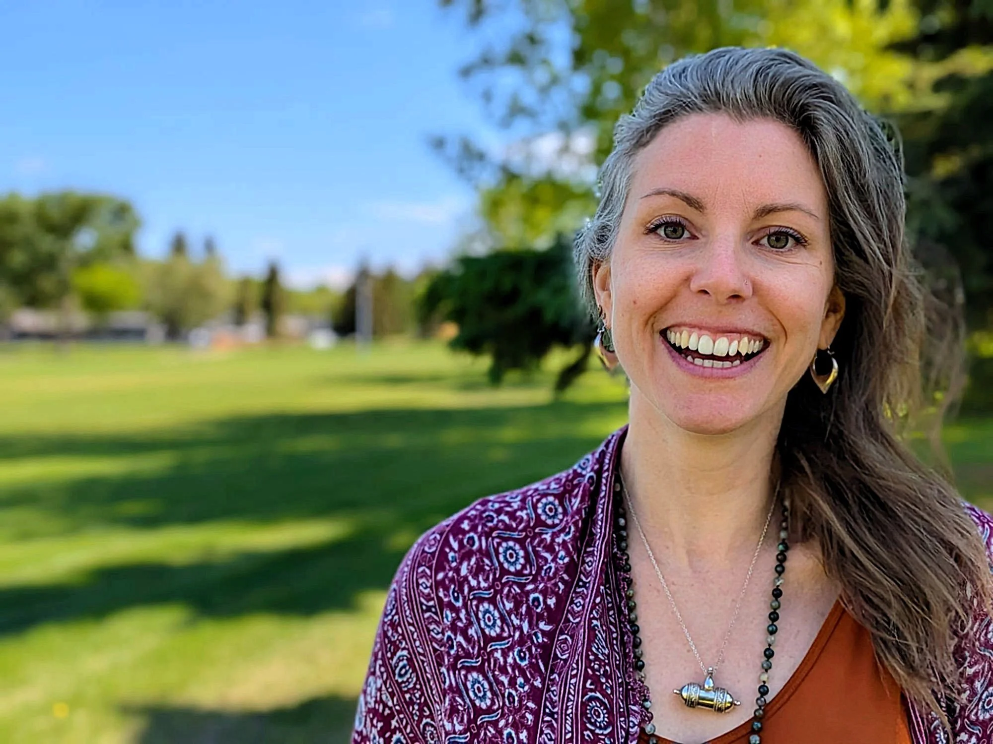 Close-up of a smiling woman with curly gray hair, wearing earrings, necklaces, and a patterned purple top, outdoors with trees and a grassy field in the background.