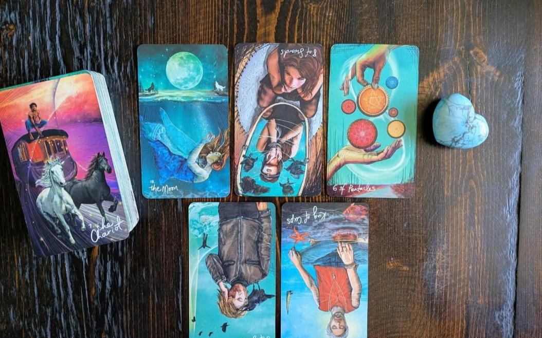 A set of tarot cards laid out on a wooden surface, with a small blue heart-shaped stone nearby.