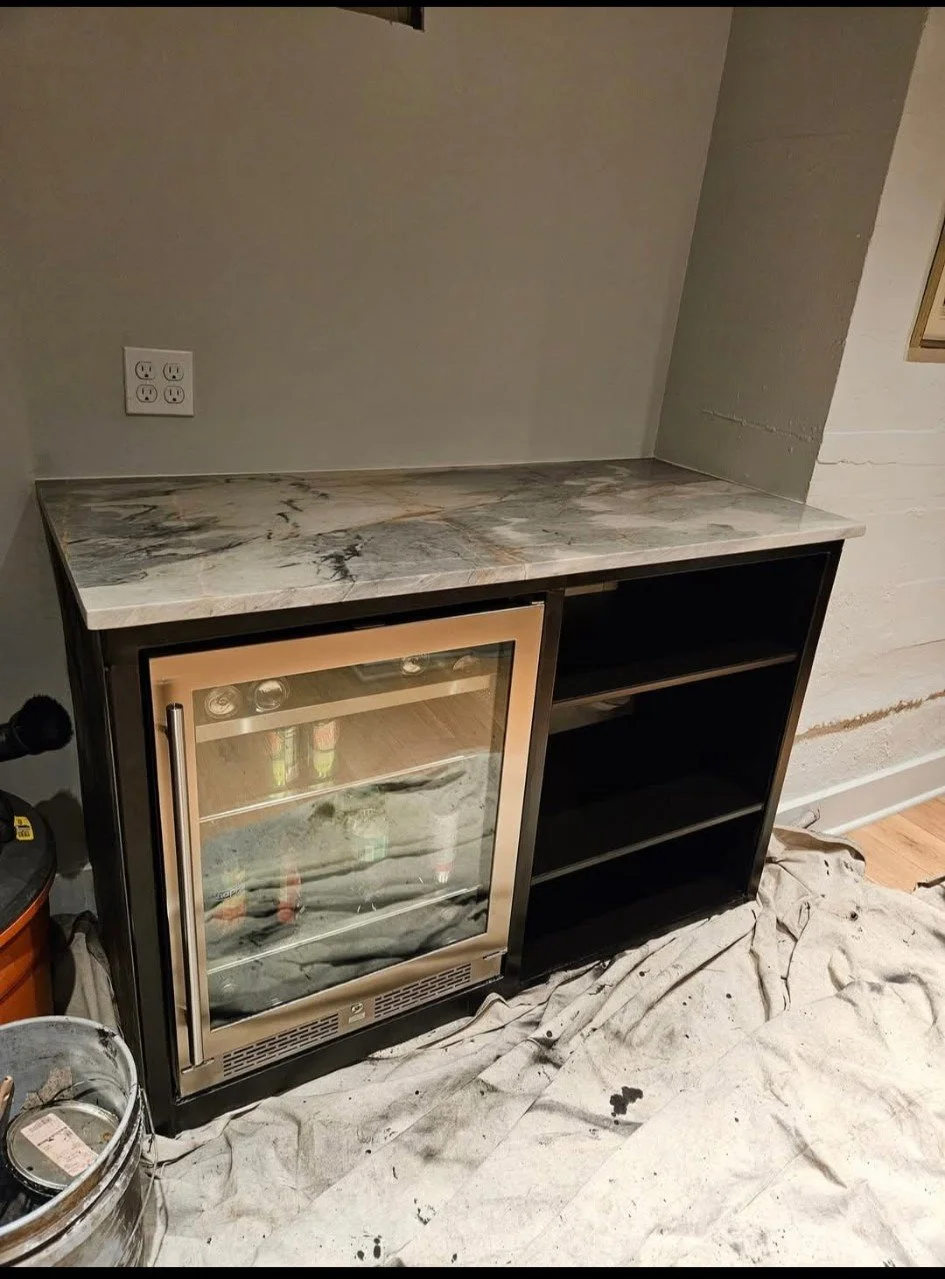 Custom modern industrial beverage center and cabinetry. Features a precision-welded, heavy-duty steel base framework that seamlessly integrates a glass-front cooler and custom shelving, topped with a premium solid-surface counter. Engineered for high
