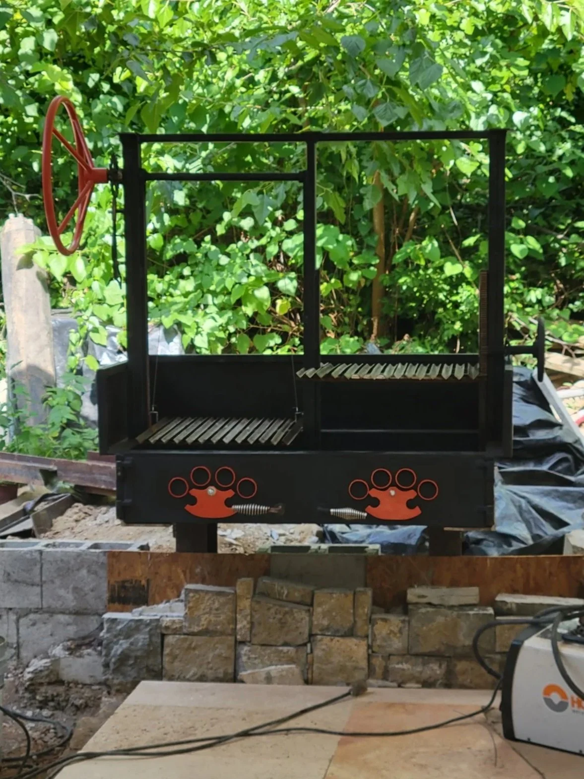 Custom-built heavy-duty charcoal grill and Santa Maria style cooker. Features an adjustable height grate with a side-crank wheel mechanism, integrated heavy steel lid, and custom heavy-duty accent plates on a durable brick-ready frame.