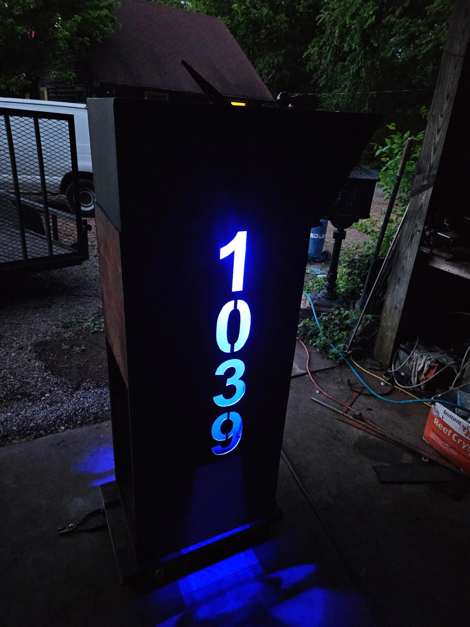 Custom modern steel mailbox post with integrated blue LED backlighting. Features precision CNC-cut vertical house numbers that glow brilliantly at night, combining striking architectural style with high-visibility curb appeal.