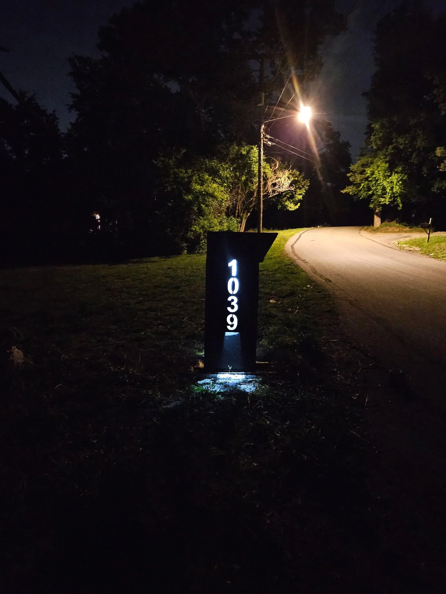 Custom architectural steel mailbox post fully installed. Showcasing its sleek modern design and high-visibility illuminated house numbers perfectly accenting the driveway entry at night.