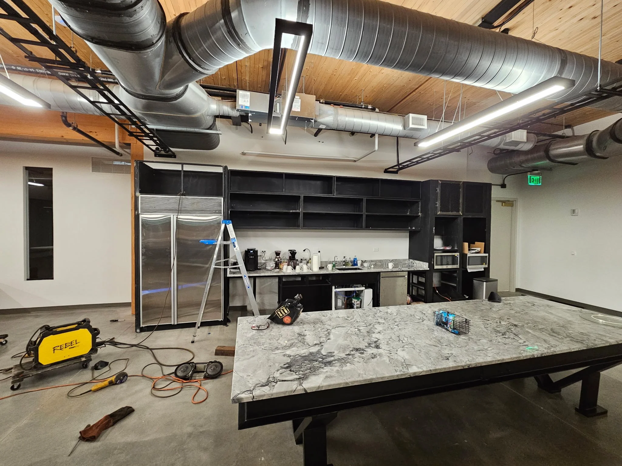 Custom commercial modern industrial kitchen build. Features a massive central island table paired with a bespoke, precision-engineered steel architectural surround housing the commercial refrigerator and integrated shelving.