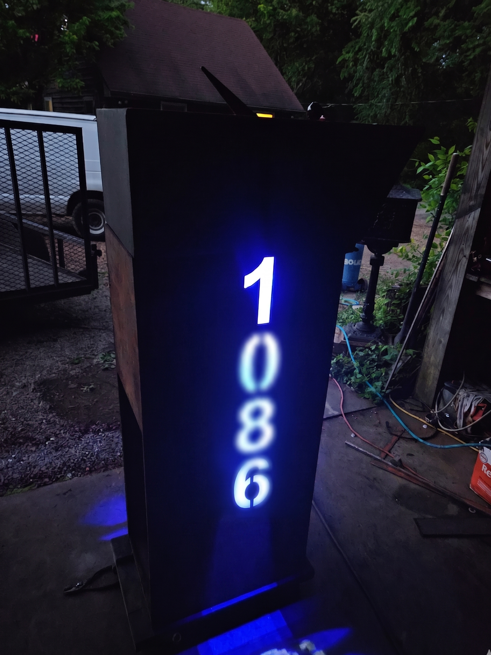 Custom modern steel mailbox post with integrated blue LED backlighting. Features precision CNC-cut vertical house numbers that glow brilliantly at night, combining striking architectural style with high-visibility curb appeal.