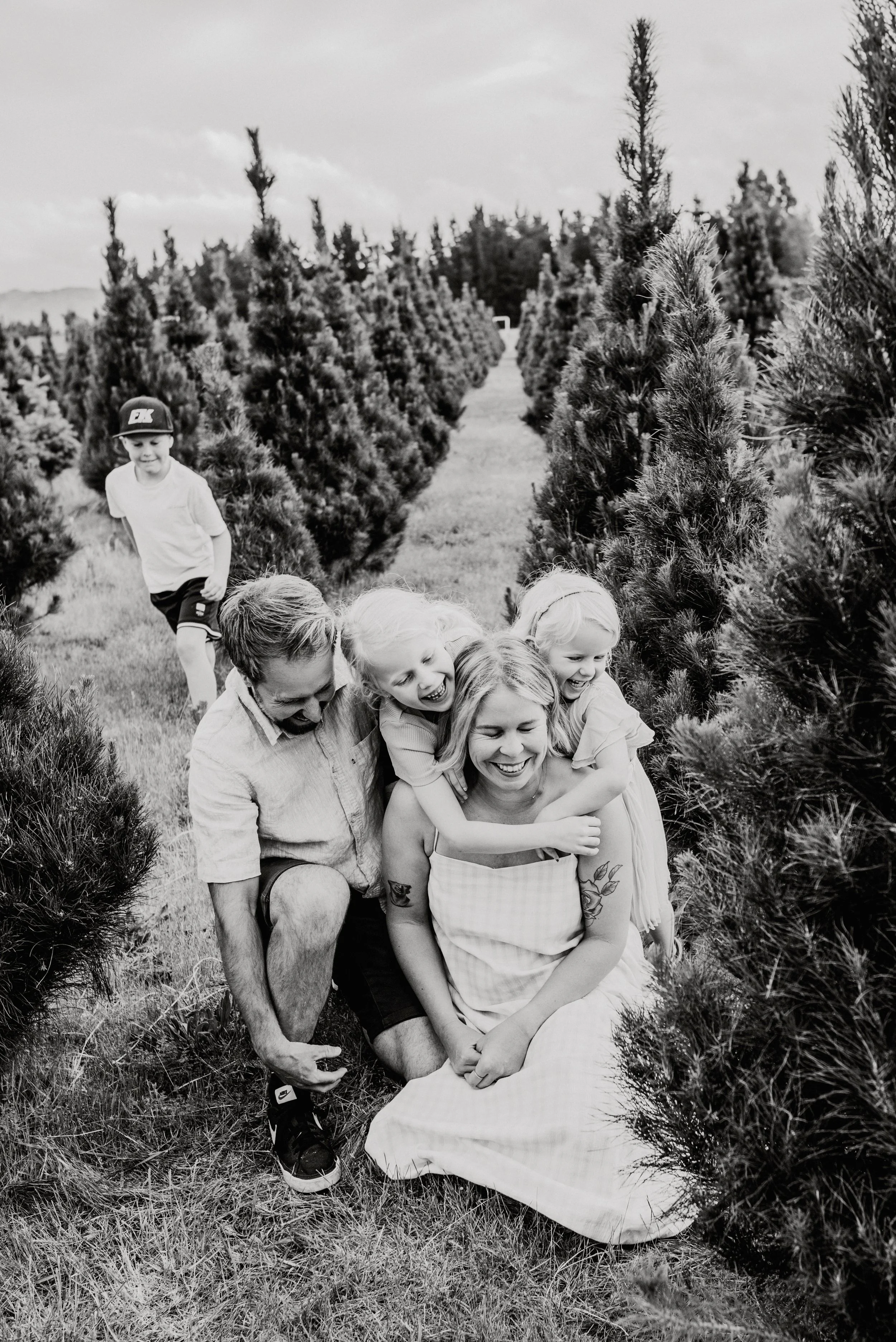 A black-and-white photo of a family of five sitting on the grass between bushes in a field, with a young boy running in the background.