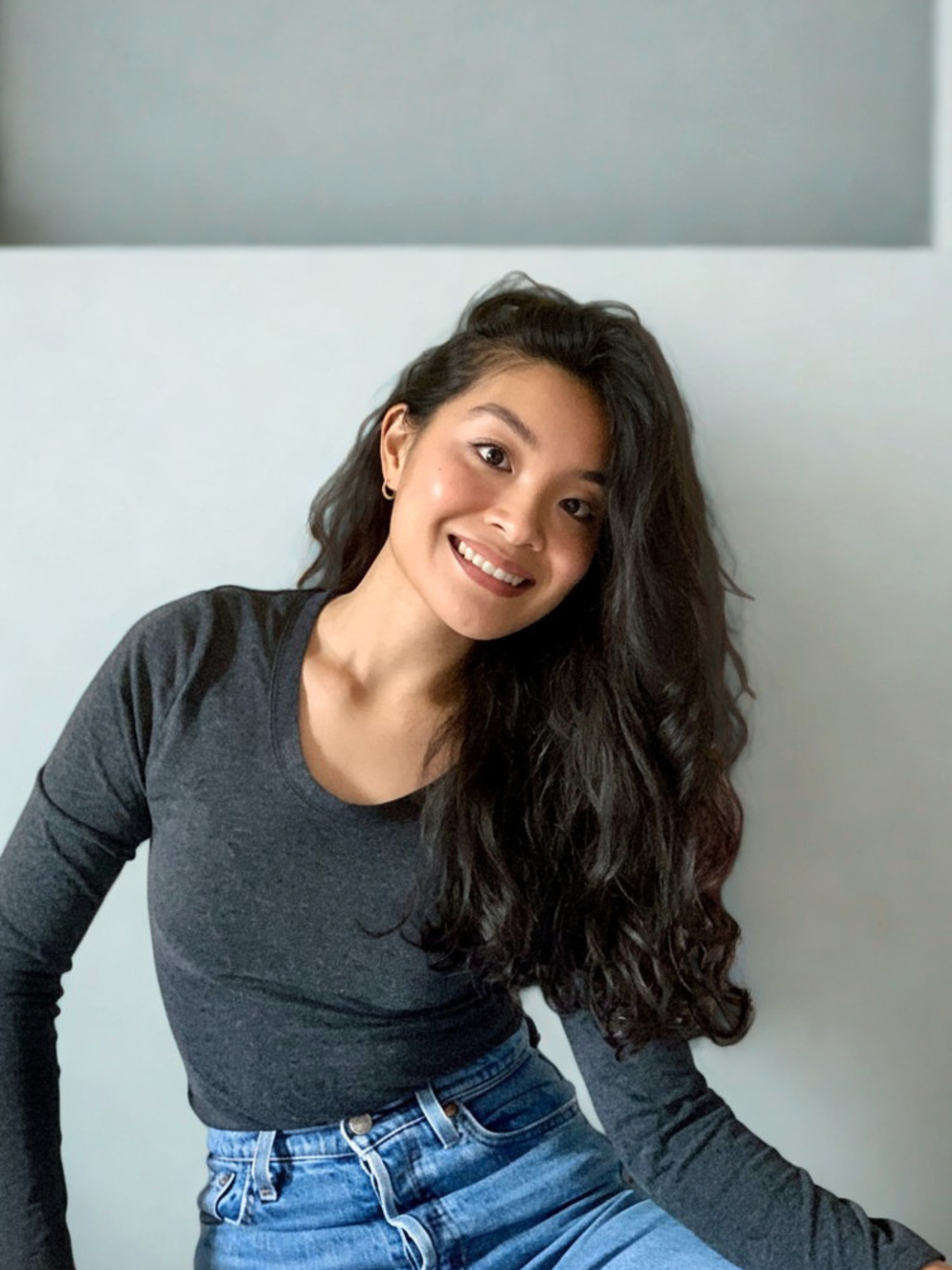 A young woman with long, wavy dark hair, smiling, wearing a dark gray long-sleeve shirt and blue jeans, sitting against a white wall.