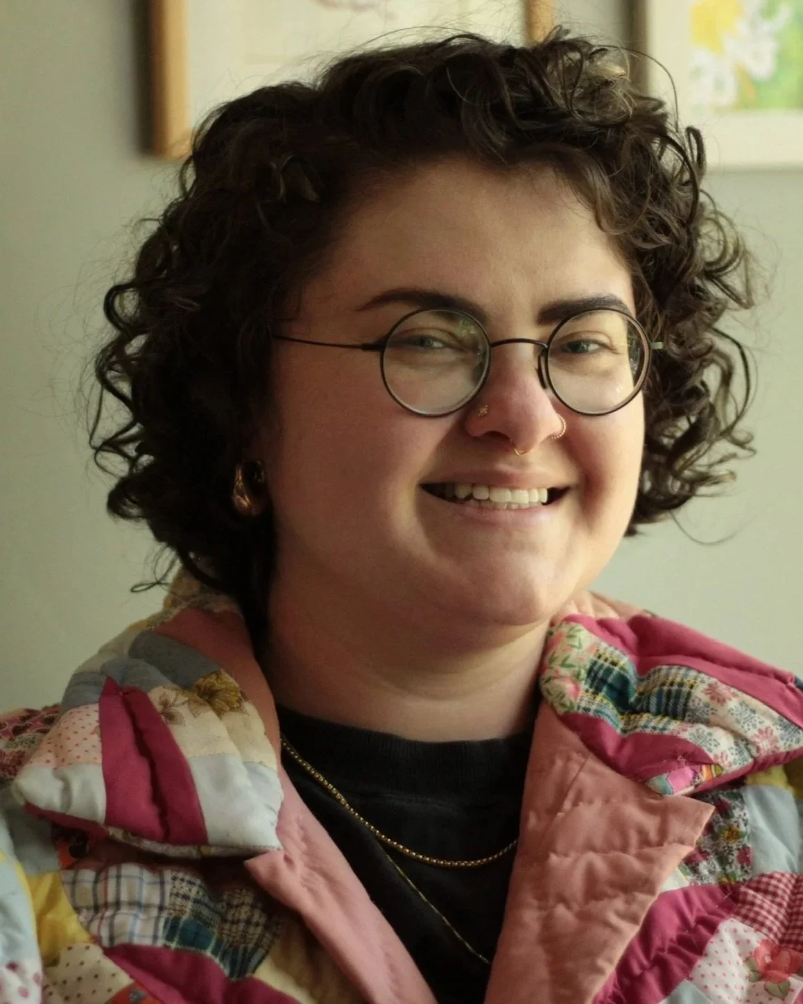 e.k., a white trans person with short curly dark hair and glasses, wearing a colorful quilted jacket and gold jewelry