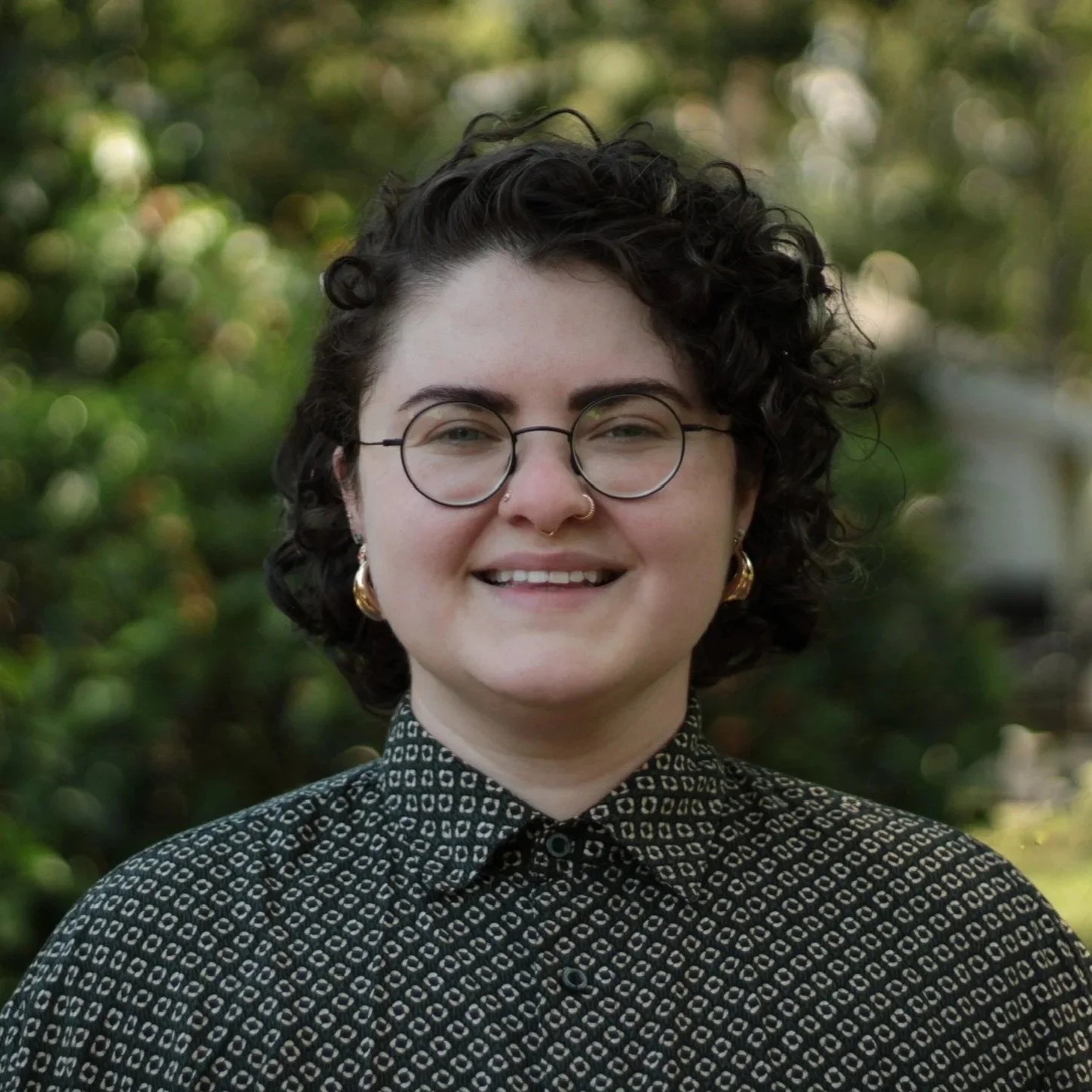e.k., a white trans person, with dark curly shoulder length hair and glasses, from the shoulders up