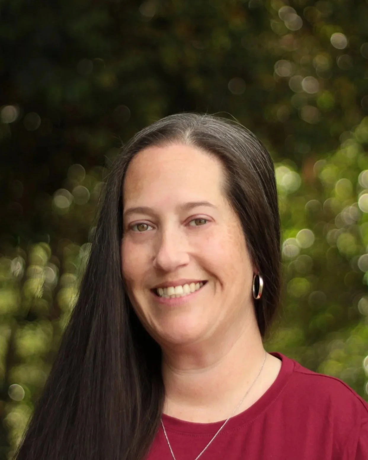 a photo of mo, a white woman with long dark straight hair, smiling with teeth, wearing a garnet shirt and silver jewelry