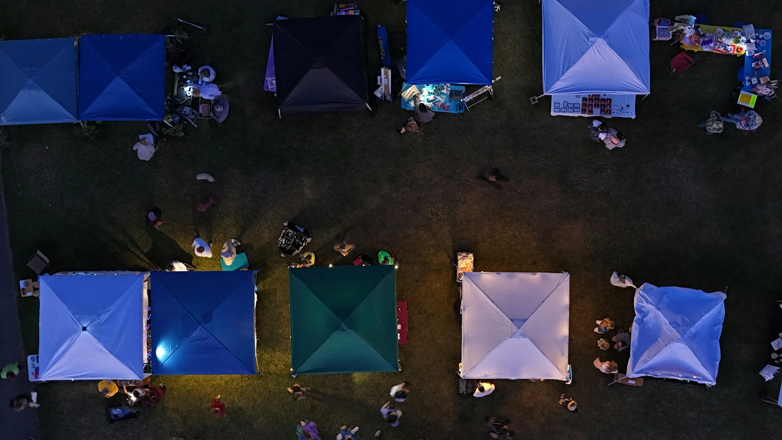Nighttime view of an outdoor market with multiple tents in shades of blue, green, purple, and white, some illuminated from within, with people browsing and shopping.