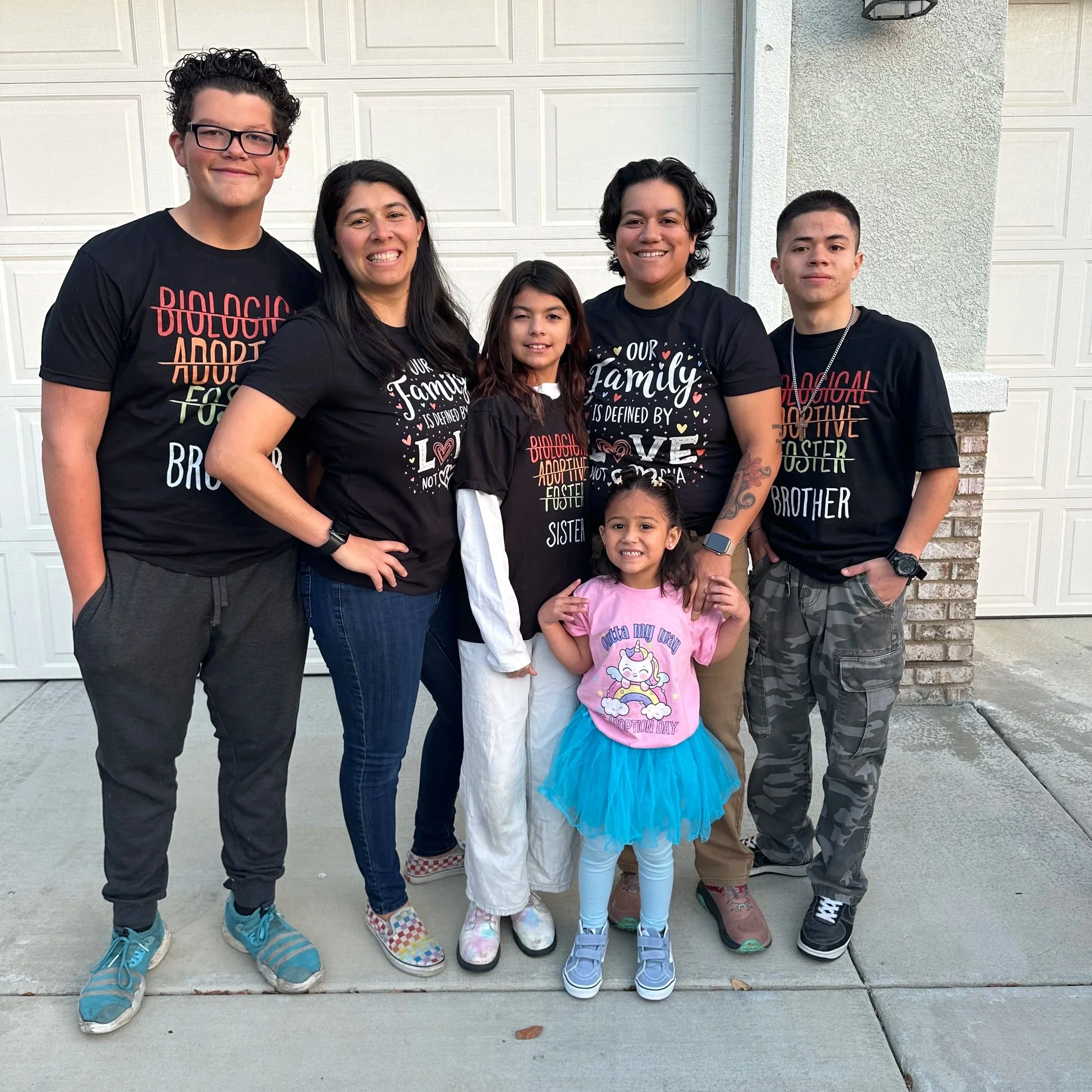 A group of seven people standing in front of a white garage door. They are all smiling and wearing black T-shirts with various messages. The group includes a tall young man with glasses, two women, a teenage boy, and three young girls. The women and girls are wearing accessories and colorful clothing.