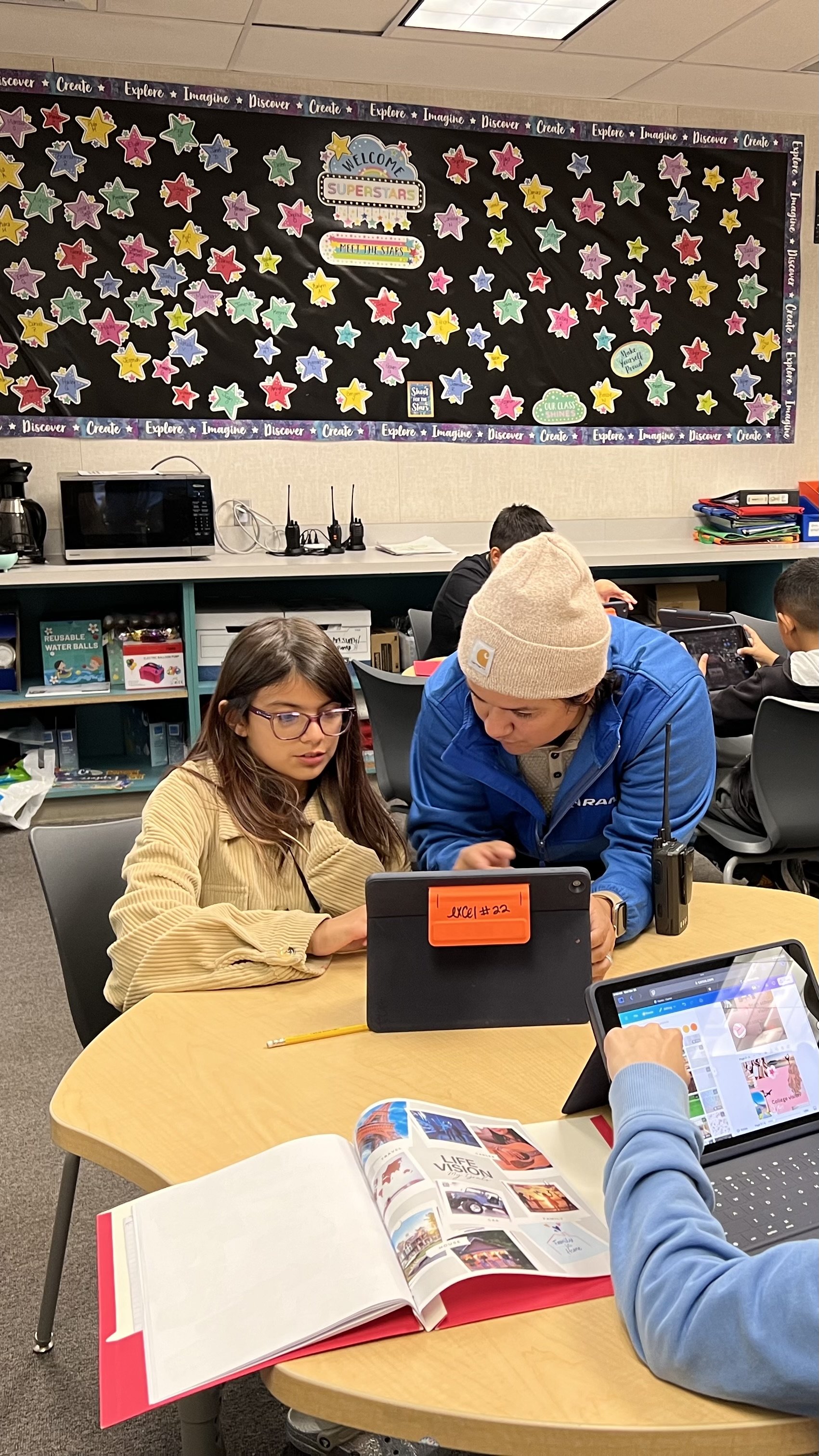 Students and teacher working together at a classroom table with books, tablets, and a laptop.