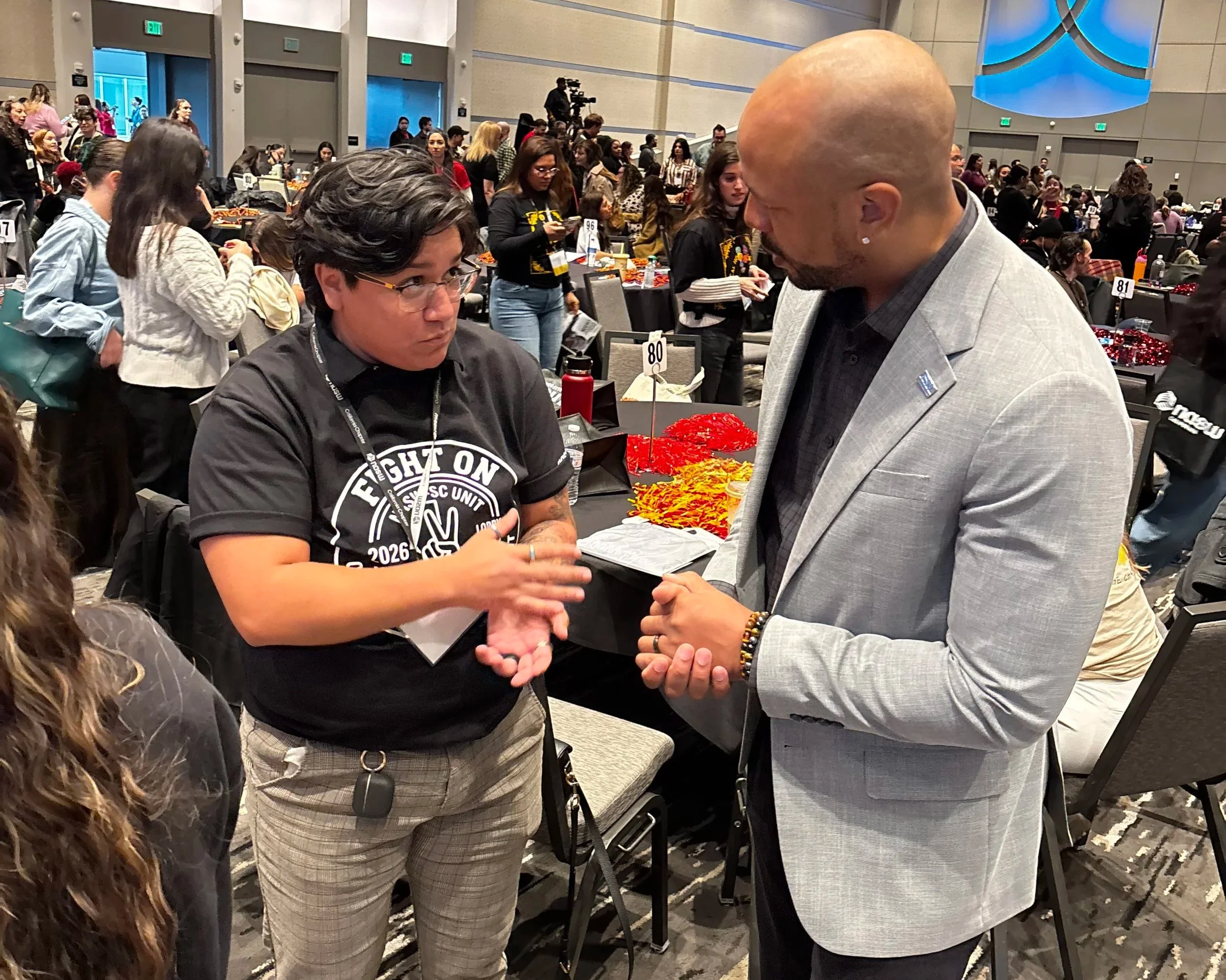 Two men are shaking hands at a conference or event, surrounded by many people sitting and standing in a large room with round tables and a high ceiling.