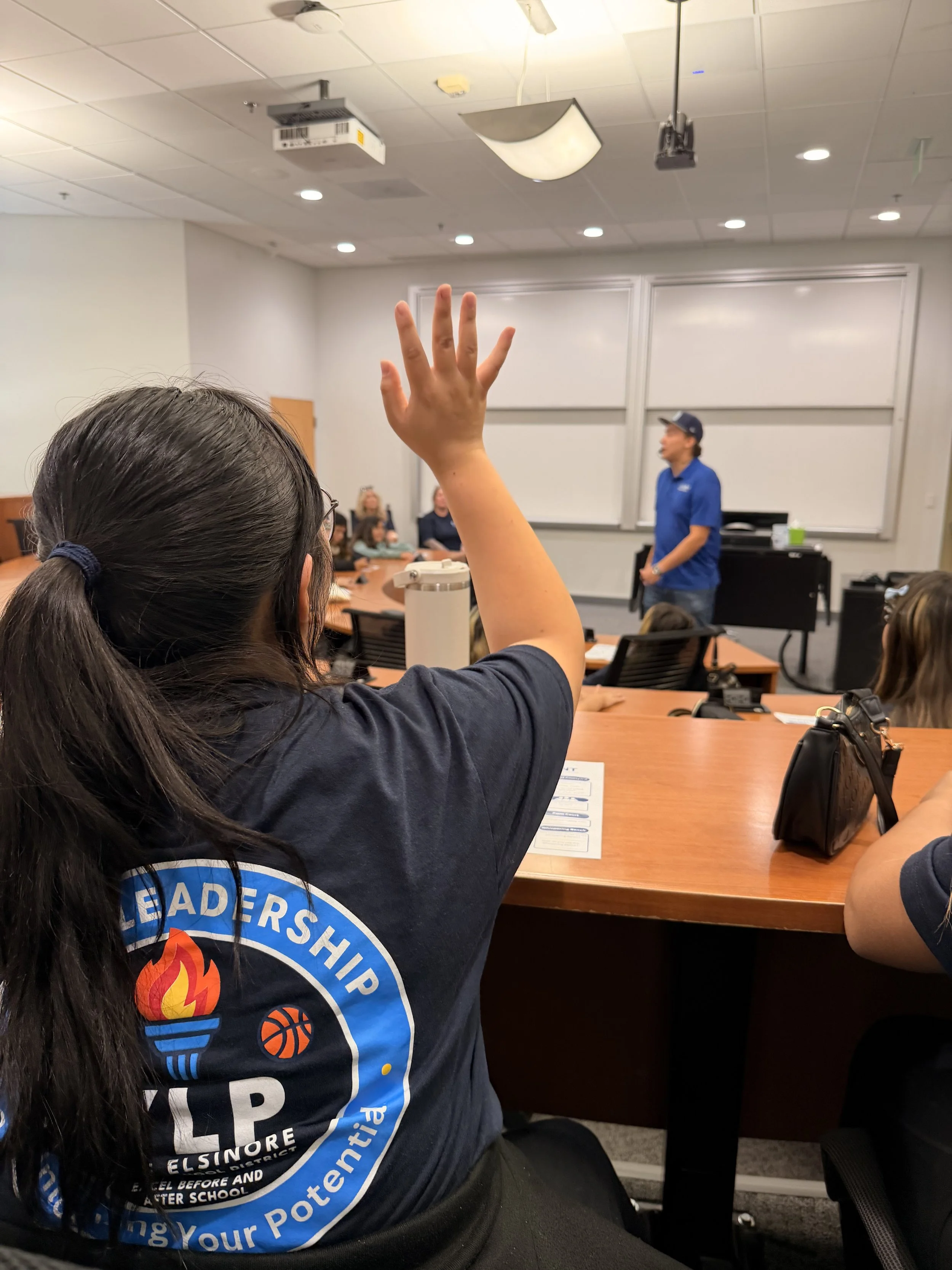 A classroom setting with a woman raising her hand. She has long, dark hair tied back and is wearing a dark T-shirt with a circular logo related to leadership, education, and youth empowerment. In the background, there is a man in a blue shirt and cap, standing near a whiteboard, with other people seated around a conference table.