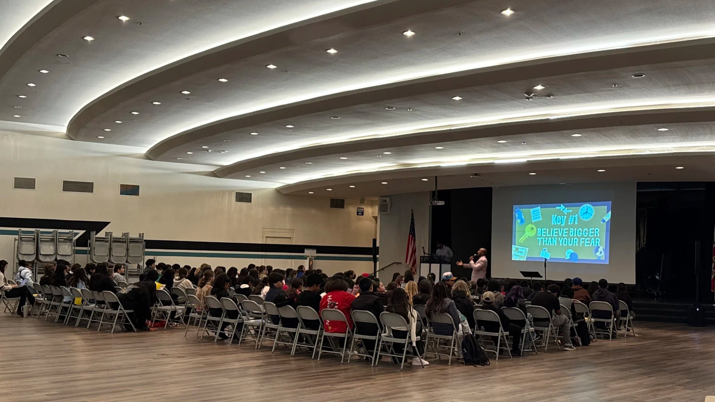 Large group of students sitting in chairs facing a stage in a school auditorium with a speaker holding a microphone. The stage has a large projection screen displaying motivational text and colorful graphics, with an American flag in the background.
