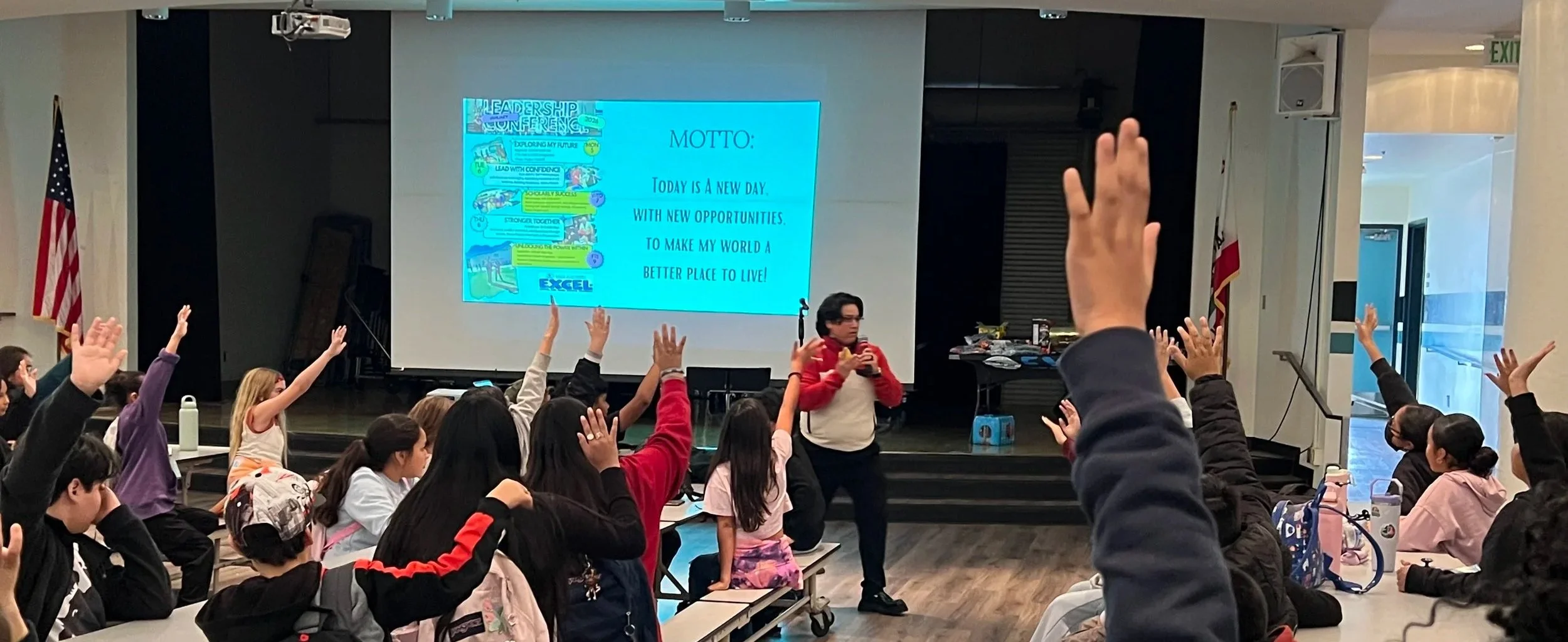 A classroom with students raising their hands during a presentation. There is a large screen displaying a blue slide with text, and a woman holding a microphone at the front.