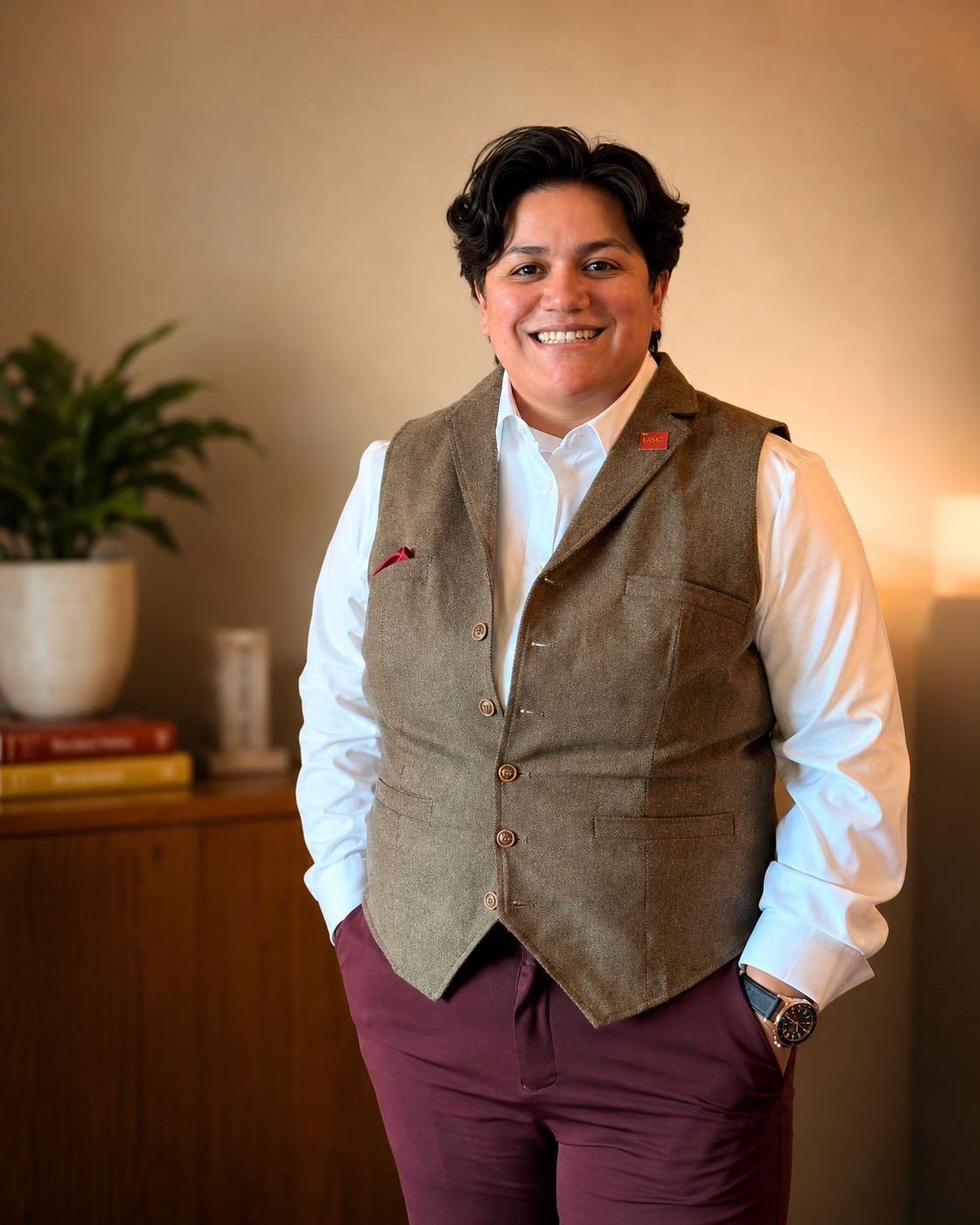 A person with short dark hair smiling, wearing a white shirt, brown vest, and burgundy pants, standing in a room with a beige wall, potted plant, and bookshelf in the background.