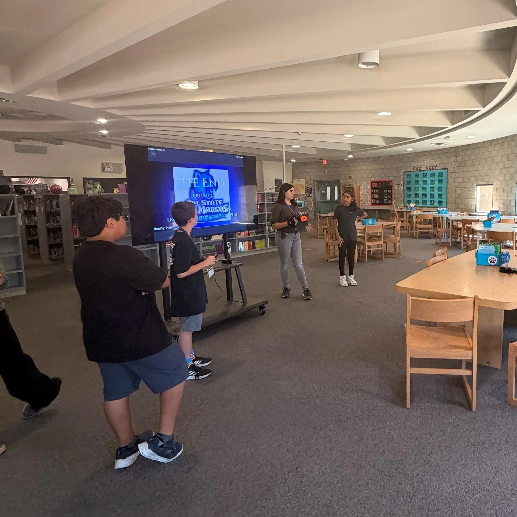 A group of children in a library or classroom, with two girls standing and speaking to a group, while three boys stand in front of her. There is a large screen displaying the words 'The End' and a classroom setting with tables and chairs.