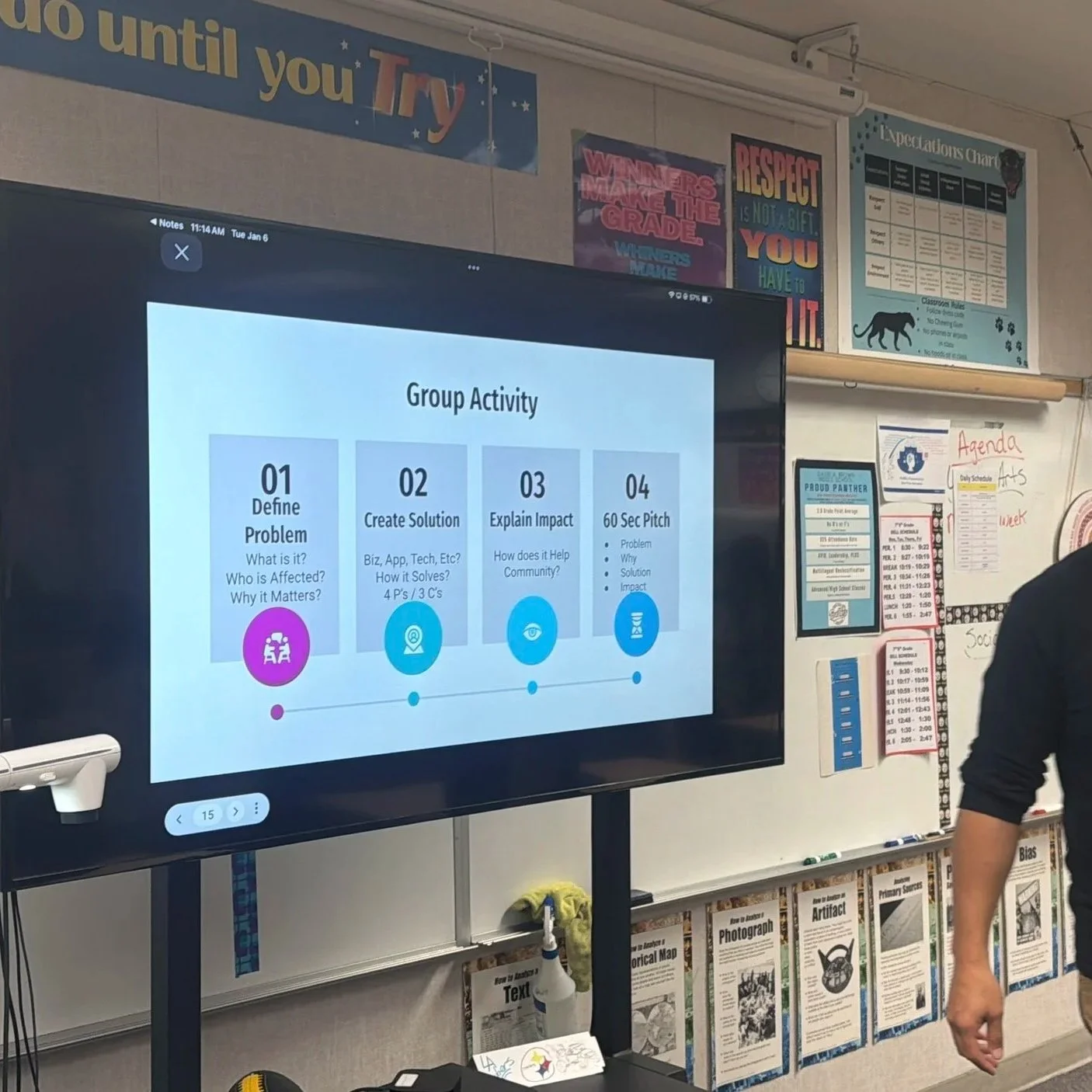 Classroom with a large digital screen displaying a group activity plan: Define Problem, Create Solution, Explain Impact, 60 Sec Pitch. The classroom walls are decorated with motivational posters, rules, and schedule outlines.