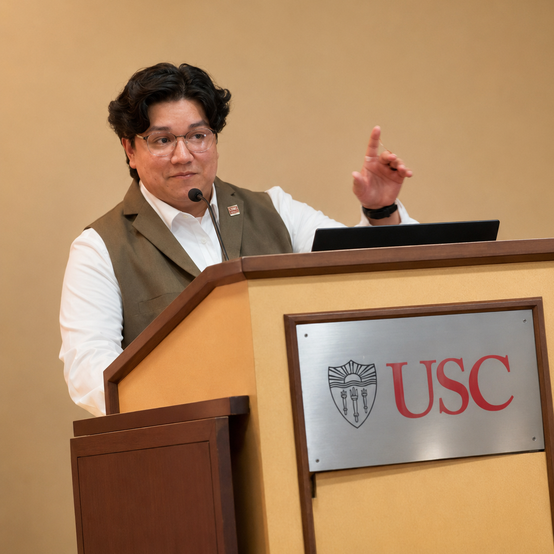 A person speaking at a podium with a USC logo, wearing glasses, a white shirt, and a brown vest.