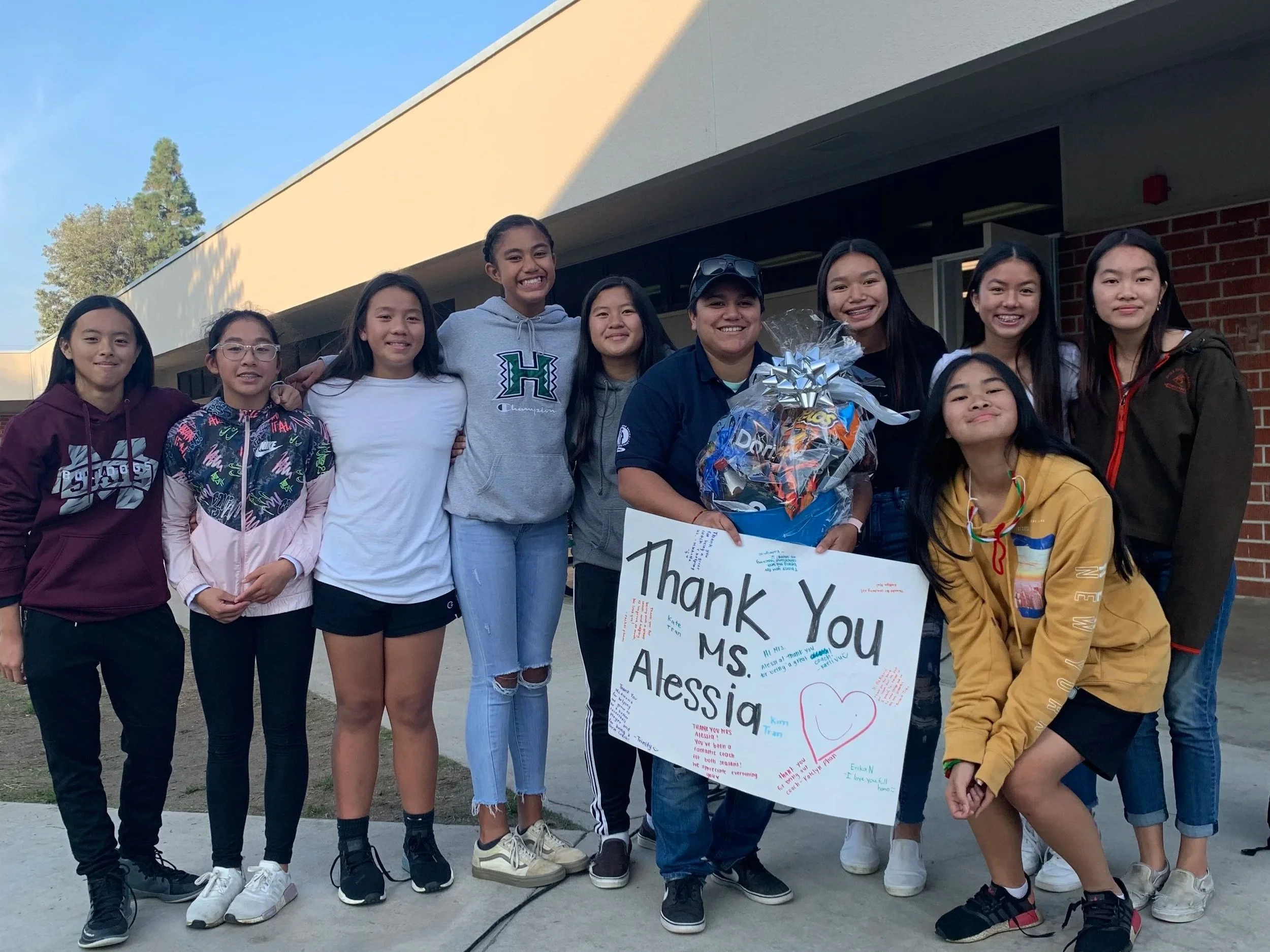 Group of ten diverse students standing outdoors, smiling, holding a large colorful thank you poster and a basket of treats, in front of a school building with trees and a light blue sky.