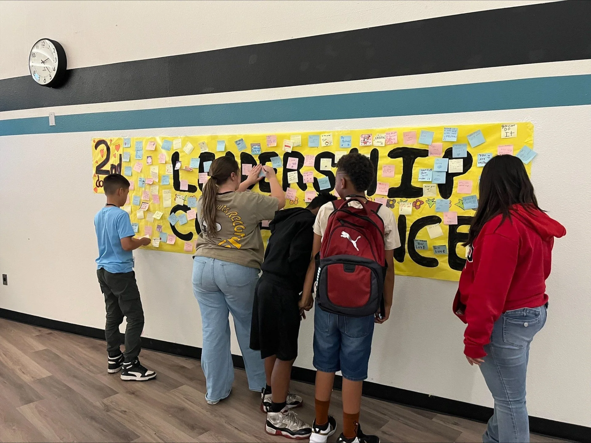 Group of children and a woman looking at a yellow bulletin board with handwritten notes and colorful sticky notes in a school hallway.