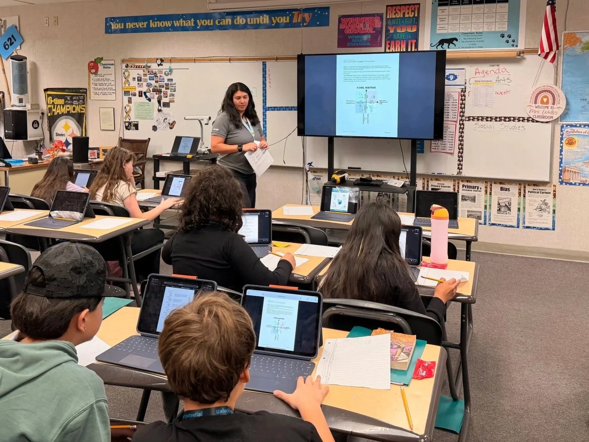 A classroom with students sitting at desks, using laptops and tablets. A woman teacher stands near a large screen displaying educational content, with posters, charts, and classroom rules on the walls.
